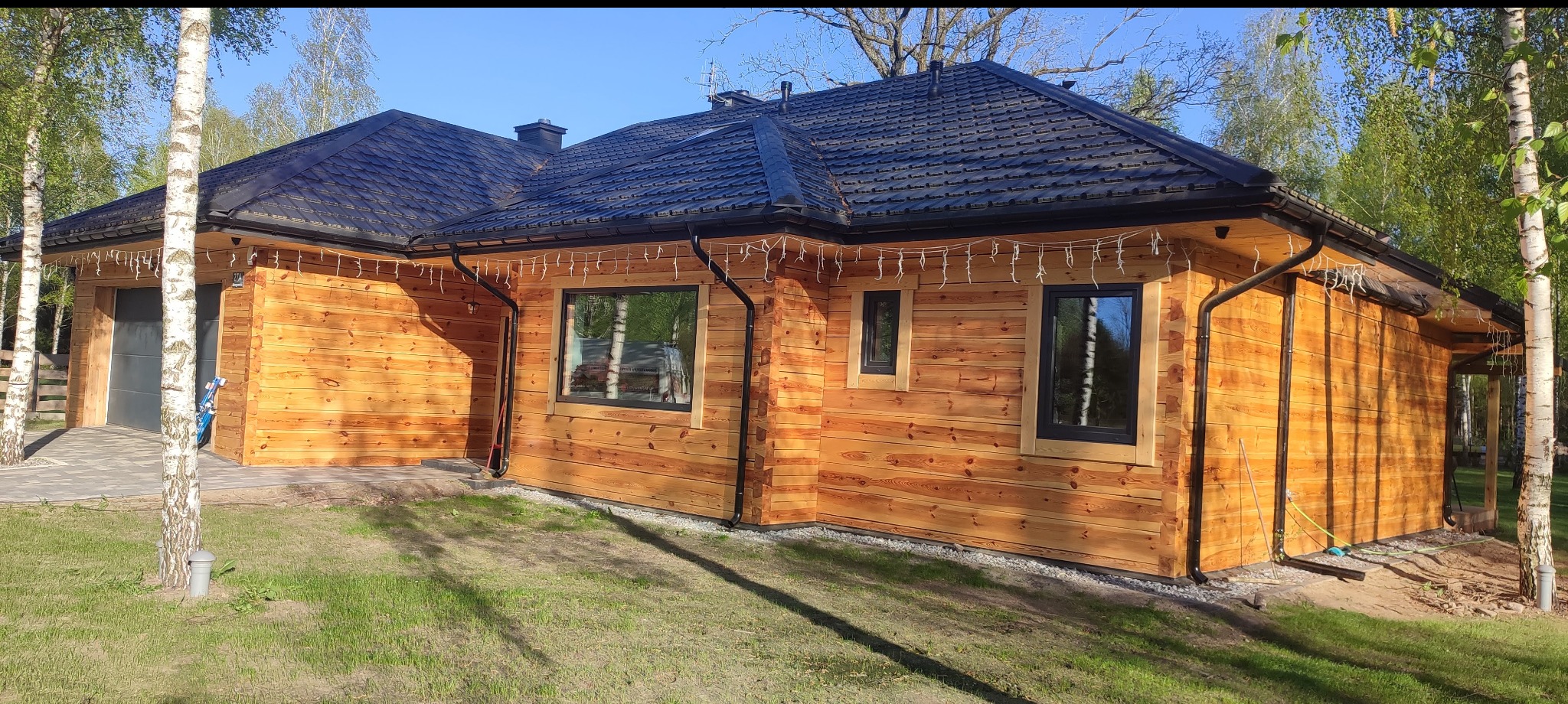 Nowoczesny dom z bali z czarnym dachem i oknami, oświetlony girlandami, otoczony zielenią. Widok z zewnątrz, podkreślający naturalne drewno i minimalistyczny design.