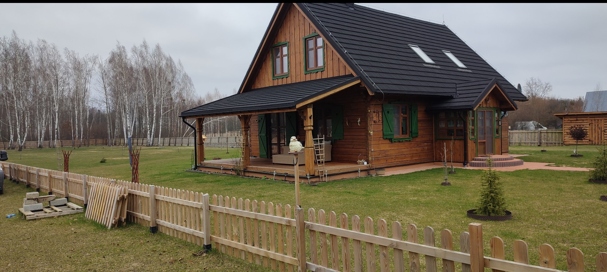 Drewniany dom z bali z czarnym dachem i zielonymi okiennicami, otoczony zielonym trawnikiem i płotem. W tle las z drzewami bez liści.
