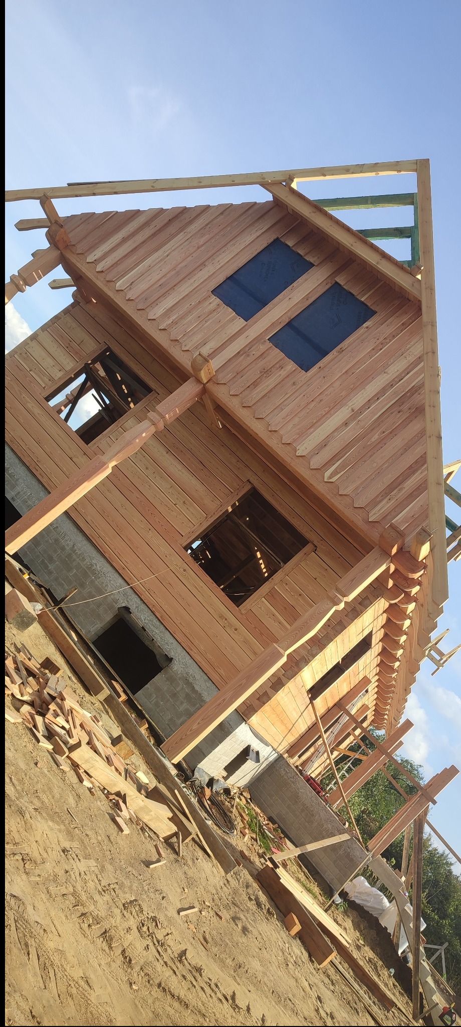 Surowy stan domu z bali w trakcie budowy, widoczne drewniane ściany, otwory okienne z niebieską folią, fundament i porozrzucane deski na placu budowy.