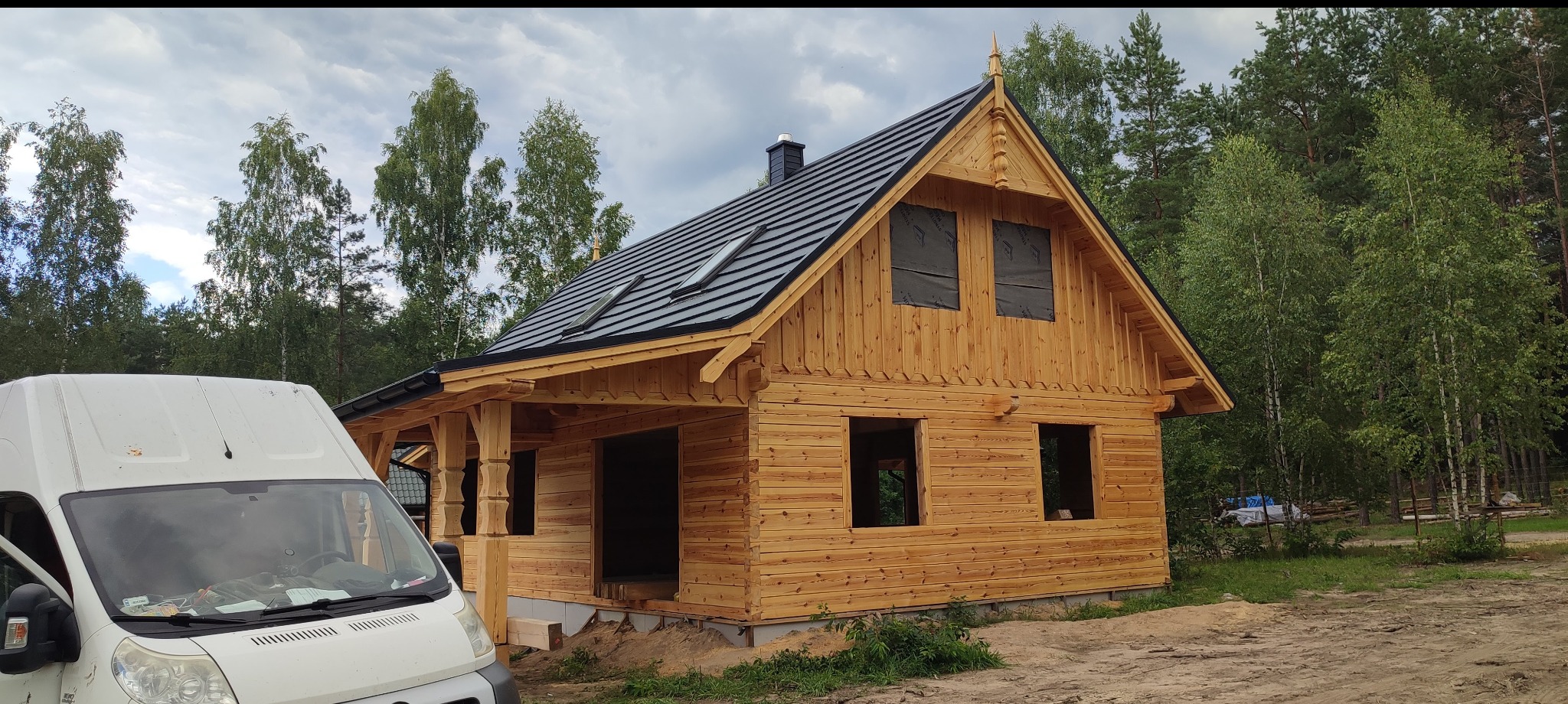 Surowy, drewniany dom z bali z czarnym dachem i oknami zabezpieczonymi folią, częściowo zasłonięty przez biały samochód dostawczy na tle lasu.
