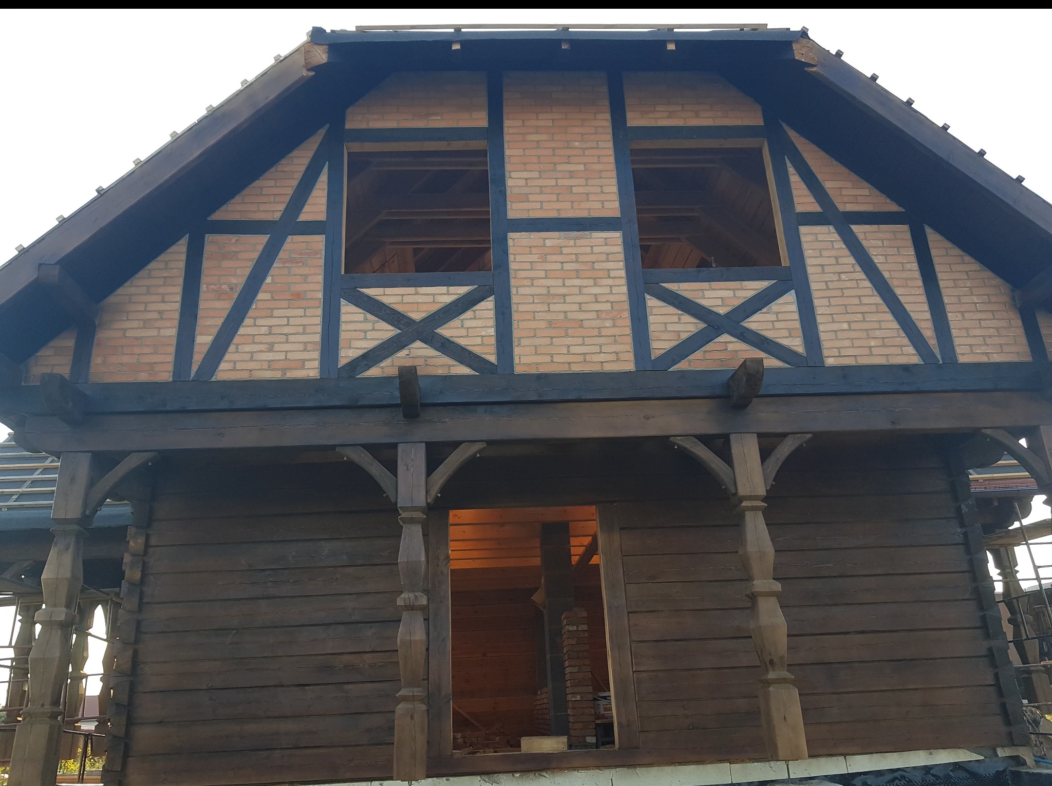 Surowy stan domu z bali z elementami muru pruskiego, widoczne niezabudowane okna i drewniane kolumny werandy. Dom w trakcie budowy, z widoczną konstrukcją dachu.