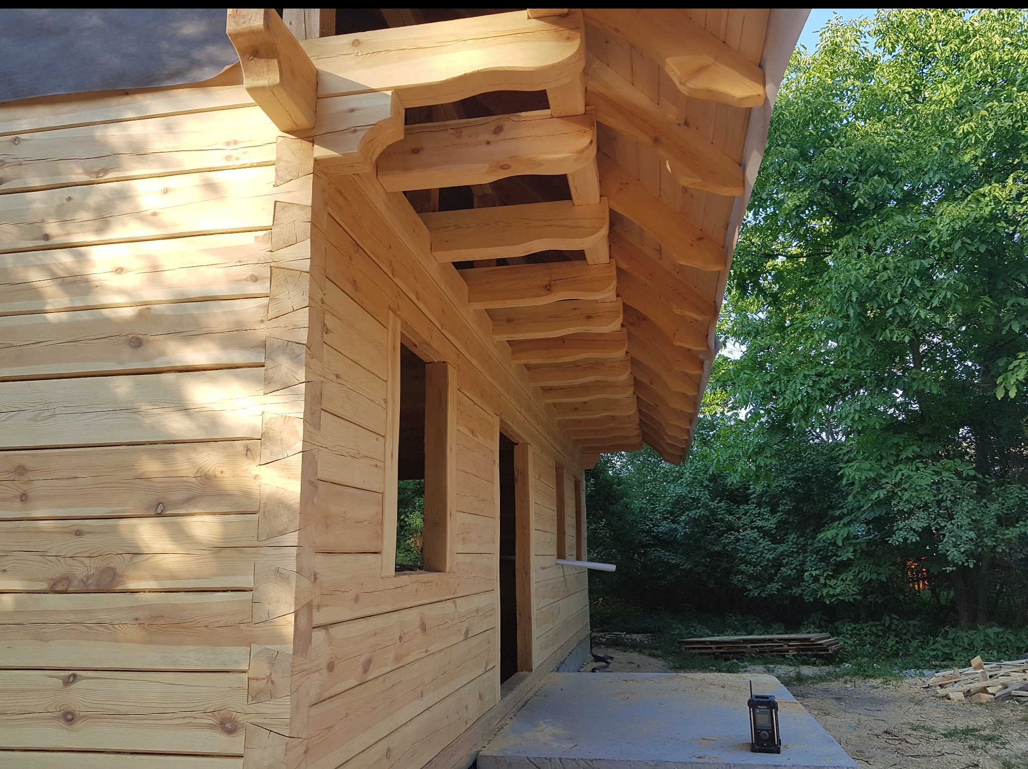 Surowy stan domu z bali w trakcie budowy, widoczne drewniane ściany, otwory okienne i ozdobne zakończenia dachu. W tle zieleń drzew.