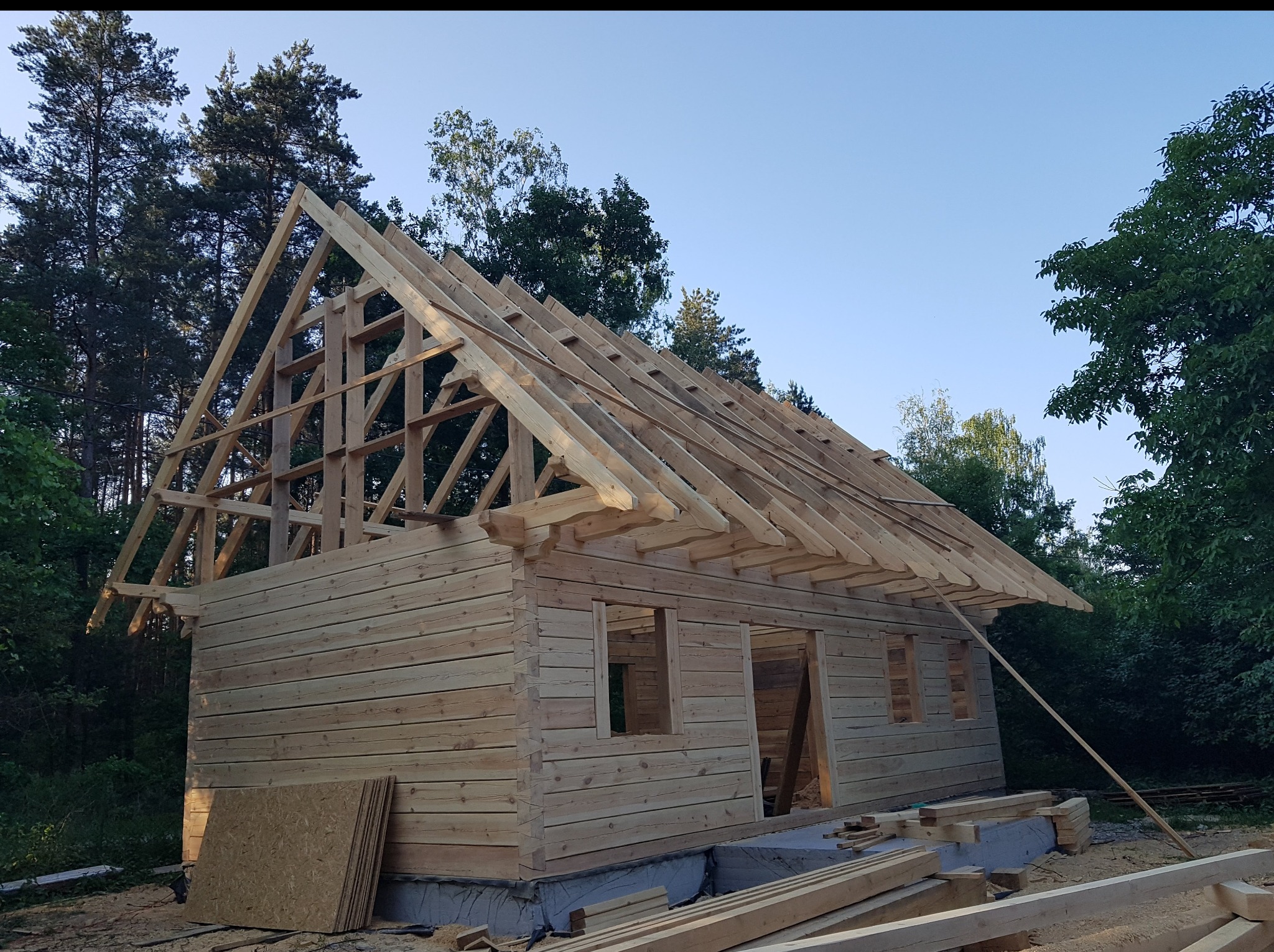 Surowy stan domu z bali w trakcie budowy, widoczne drewniane ściany, konstrukcja dachu i elementy wykończeniowe na tle zieleni i błękitnego nieba.