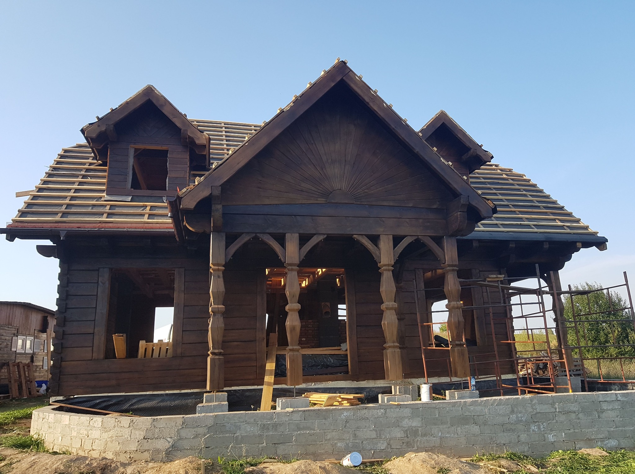 Surowy, ciemnobrązowy dom z bali w trakcie budowy, z widoczną konstrukcją dachu i fundamentami z cegieł. Charakterystyczne kolumny wejściowe i dekoracyjne elementy drewniane.