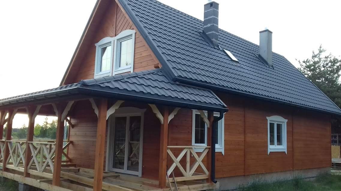 Drewniany dom z bali z ciemnym dachem i werandą z ozdobnymi balustradami. Widoczne detale wykończenia i komin. Ujęcie z zewnątrz, w naturalnym oświetleniu.