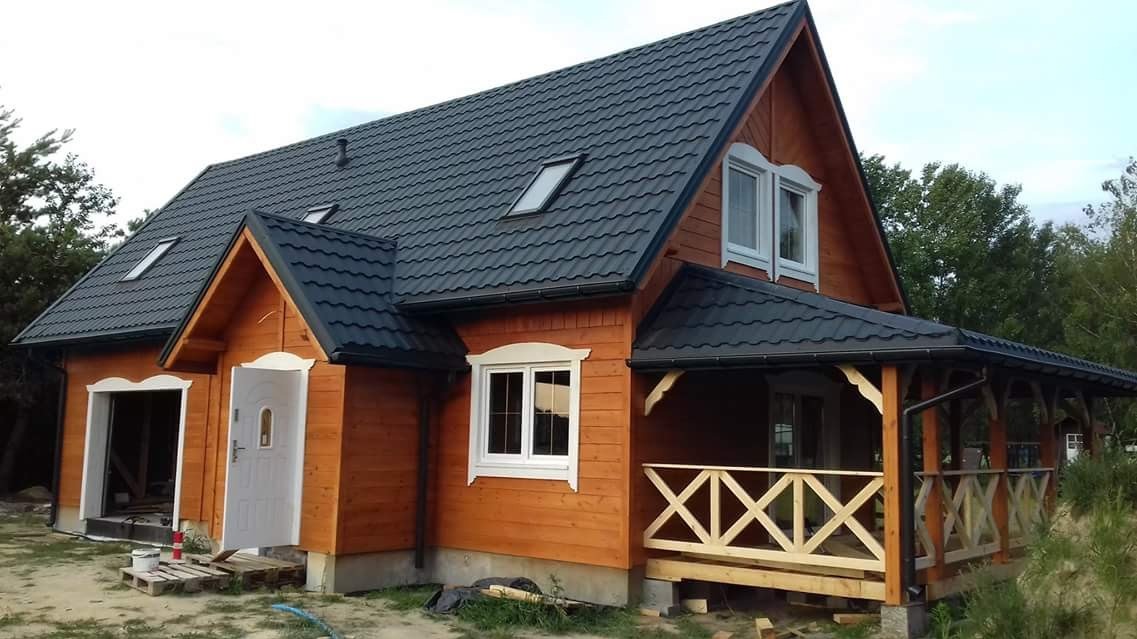 Nowoczesny dom z bali z ciemnym dachem i jasną stolarką okienną. Widoczny zadaszony taras i otwarty garaż. Drewniana elewacja w ciepłym odcieniu brązu.