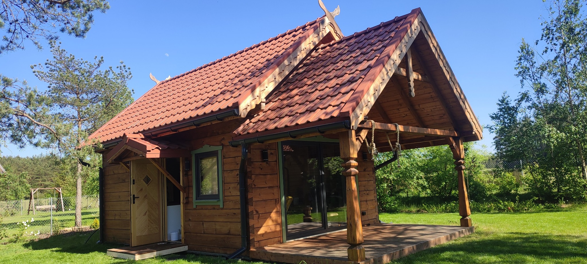 Urokliwy, parterowy dom z bali z czerwonym dachem w Nowogrodzie. Widoczna weranda z drewnianymi kolumnami i dekoracyjnymi detalami. Jasne, słoneczne otoczenie.