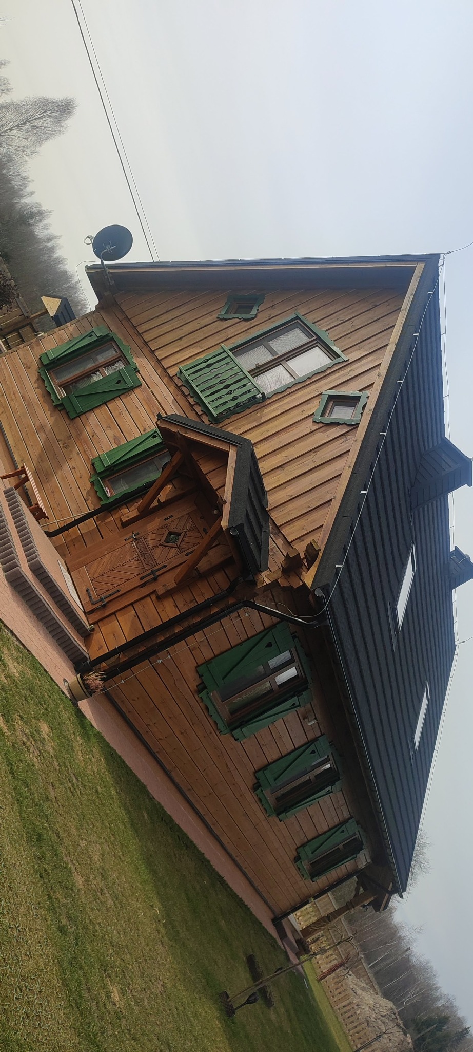 Nietypowy dom z bali z zielonymi okiennicami i ciemnym dachem, widziany pod kątem. Na dachu antena satelitarna. Otoczenie: trawnik i drzewa.