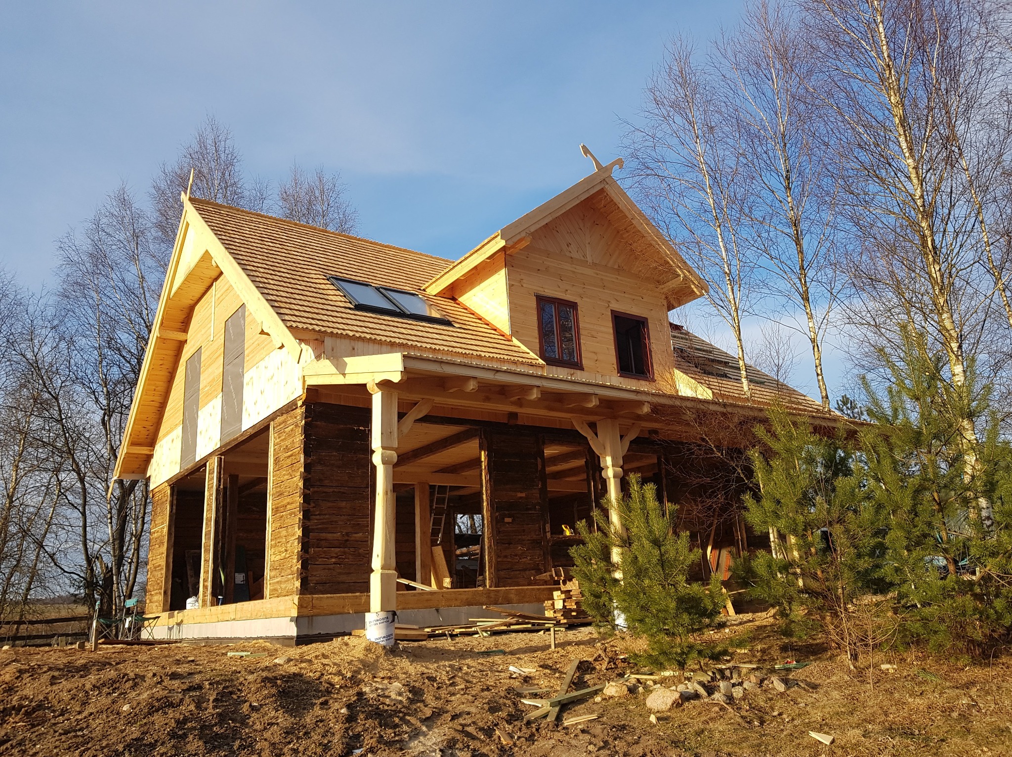 Surowy stan domu z bali z drewnianym dachem i oknami dachowymi, widoczne elementy konstrukcyjne i otwarte przestrzenie, otoczenie naturalne z drzewami.