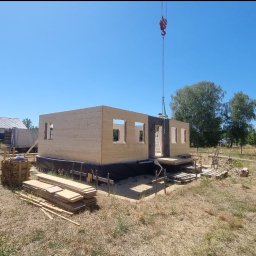 DomDrewniane DK Damian Stępień - Montaż segmentu domu modułowego z drewna na fundamencie, widoczny dźwig i transport materiałów. Słoneczny dzień, budowa w toku.