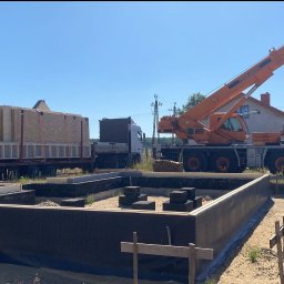 DomDrewniane DK Damian Stępień - Montaż domu modułowego: dźwig unosi element konstrukcyjny nad przygotowanym fundamentem. Widoczny transport z elementami domu. Słoneczny dzień.