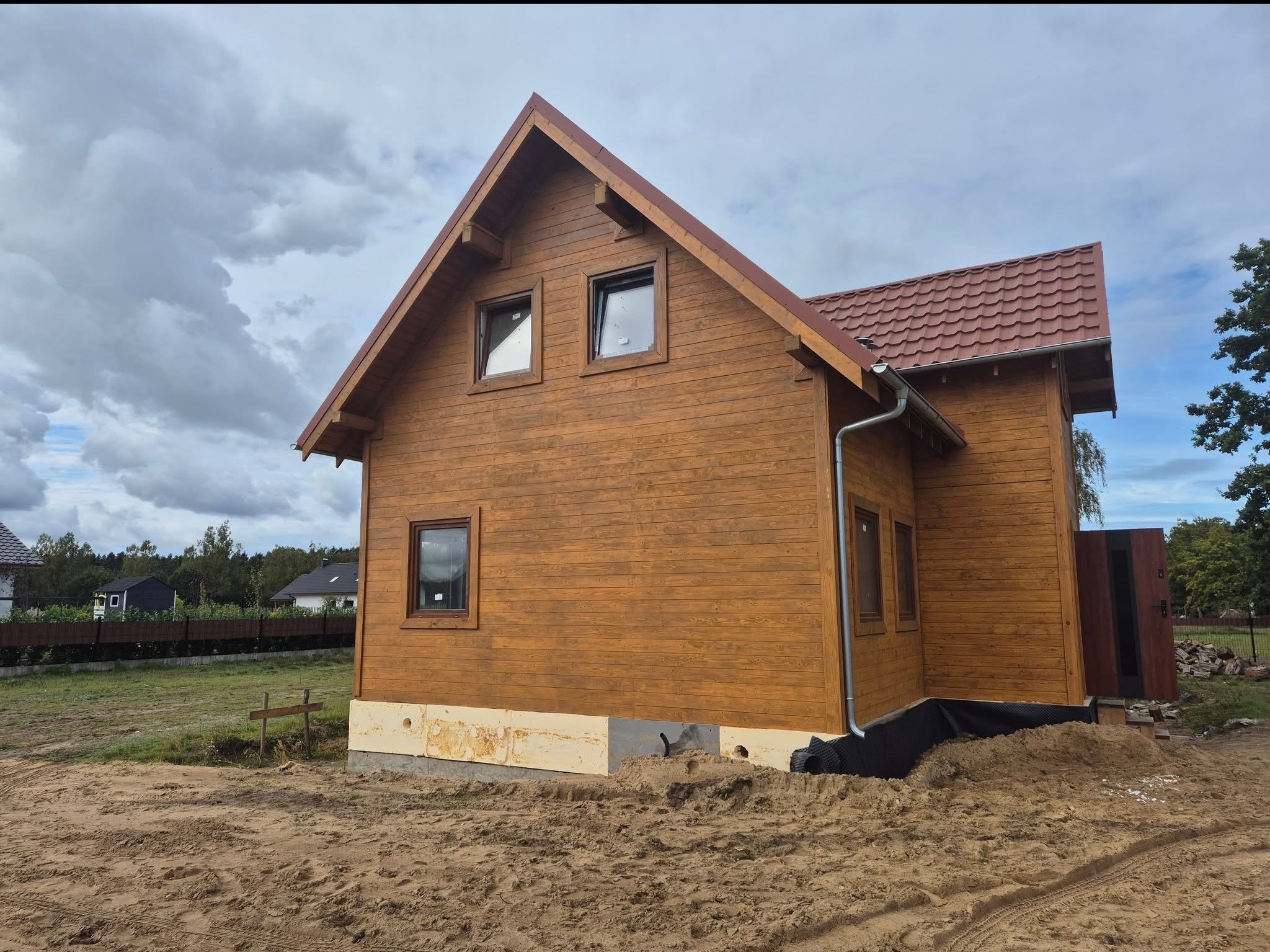 Drewniany dom z czerwonym dachem w trakcie budowy, widoczne ocieplone fundamenty i piasek. Ujęcie z boku, pochmurne niebo w tle.