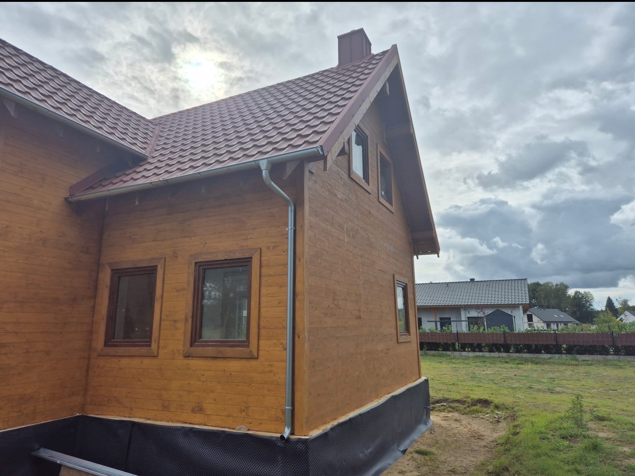 Wykończony dom szkieletowy z brązową elewacją i dachem z dachówki, widoczny fragment fundamentu i orynnowanie. W tle inne budynki i pochmurne niebo.
