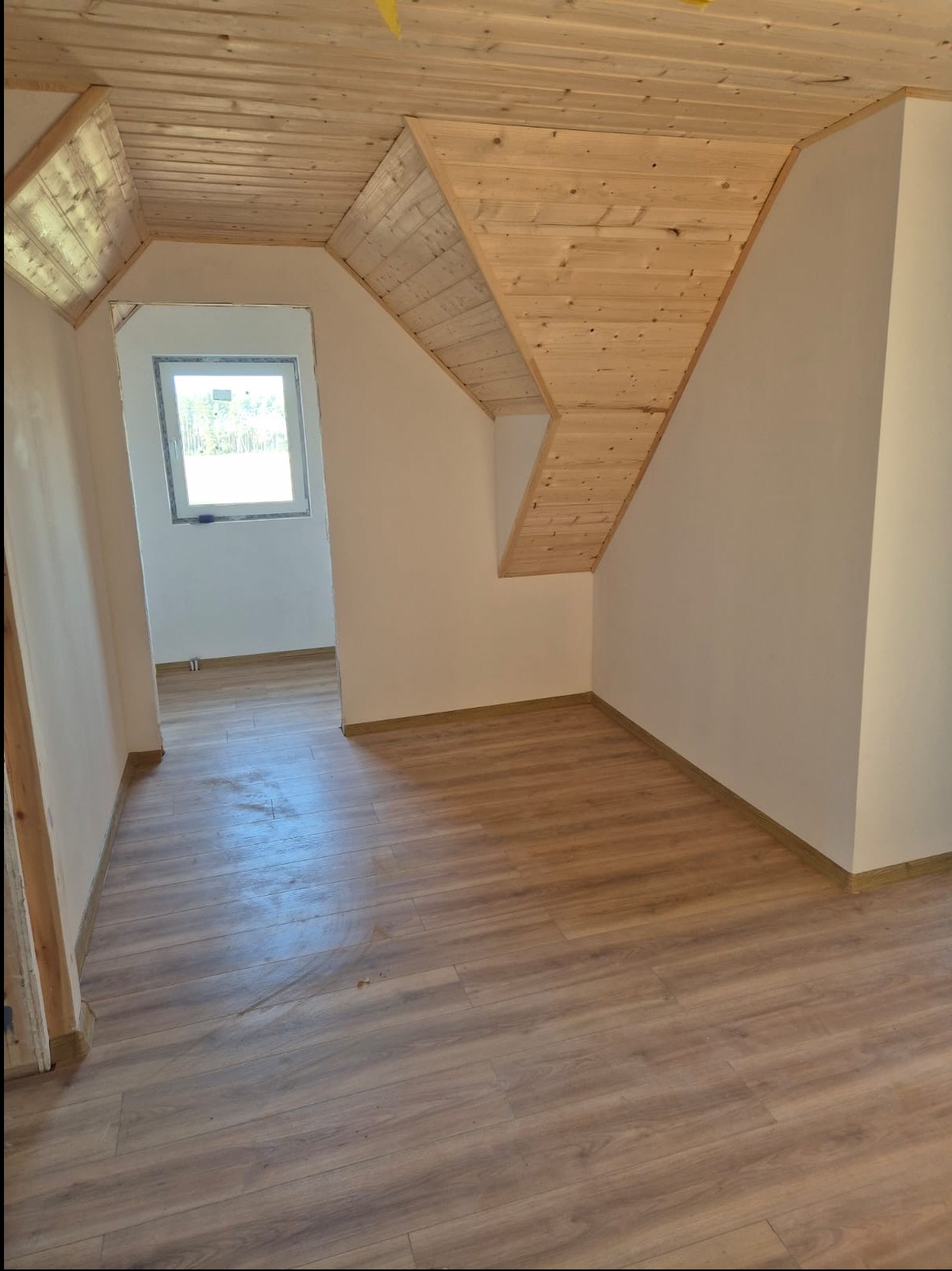Wnętrze poddasza z drewnianą boazerią na skośnym suficie i panelami podłogowymi. Widoczne okno i fragment korytarza. Ściany pomalowane na jasny kolor.