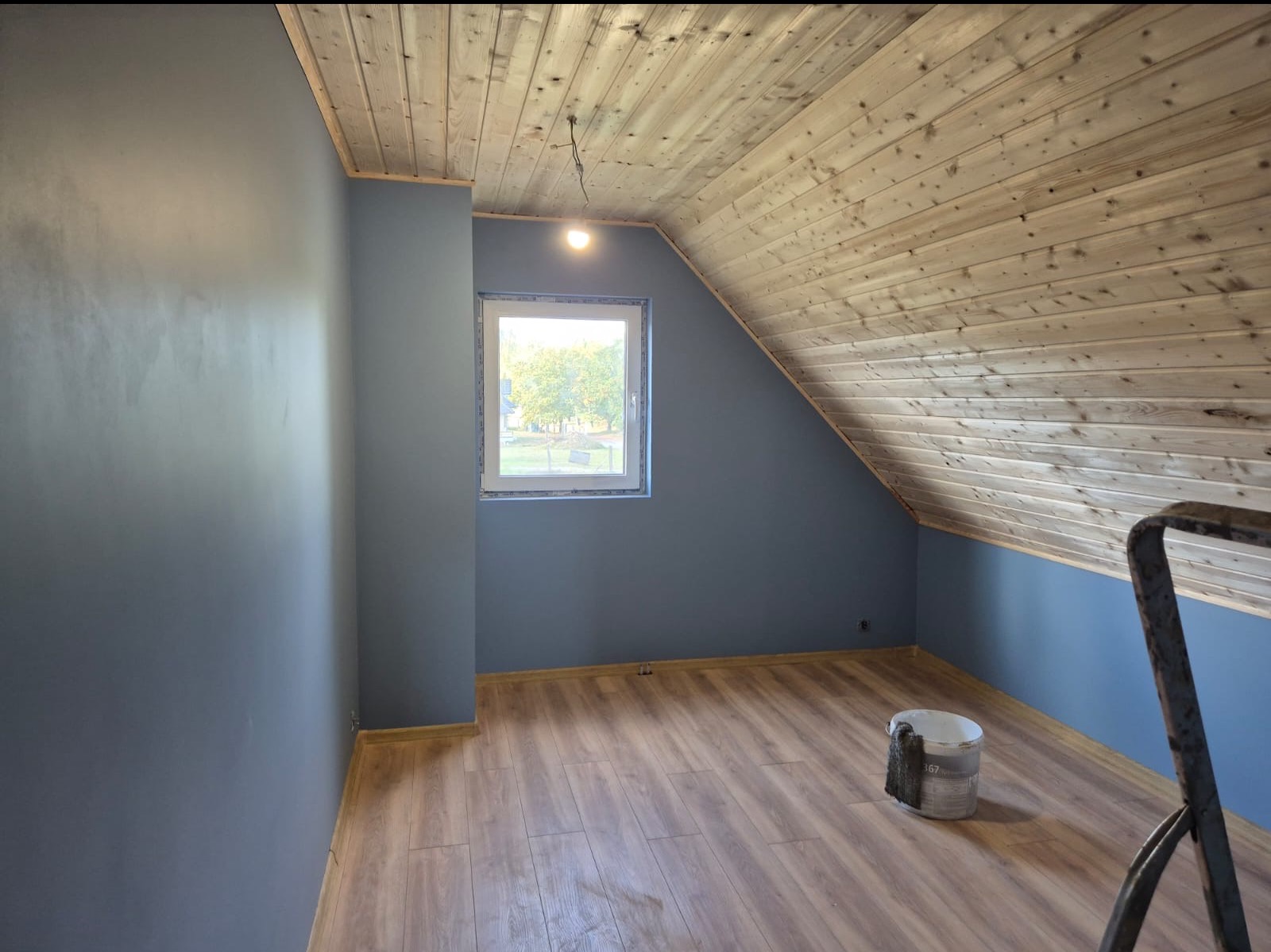 Pokój na poddaszu po malowaniu ścian na szaro, z drewnianą boazerią na suficie i oknem. Na podłodze panele, widoczne wiadro z farbą i fragment drabiny.