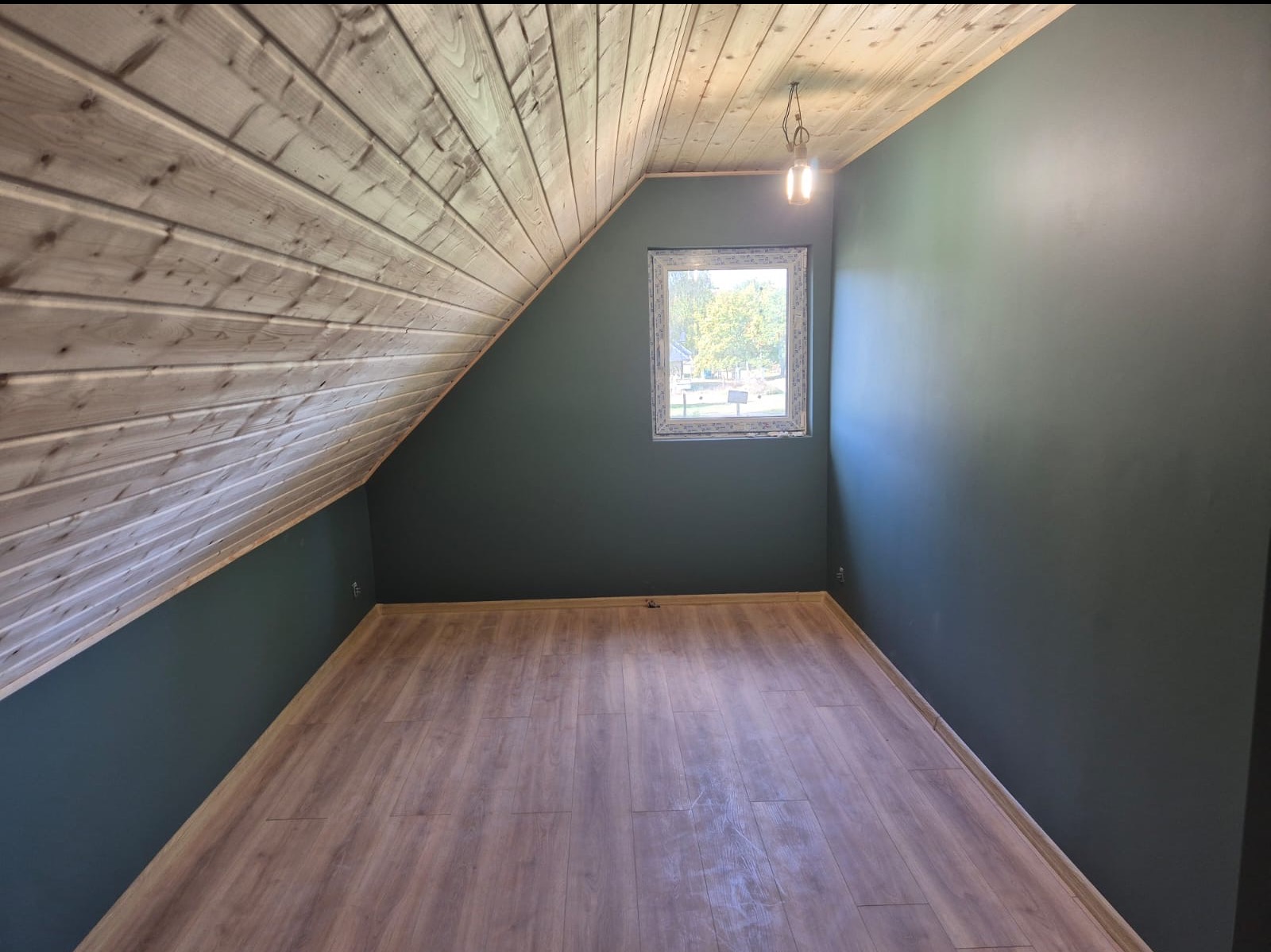 Pokój na poddaszu z drewnianą boazerią na skośnym suficie, pomalowanymi na ciemnozielono ścianami i panelami podłogowymi. Widoczne okno i wisząca żarówka.