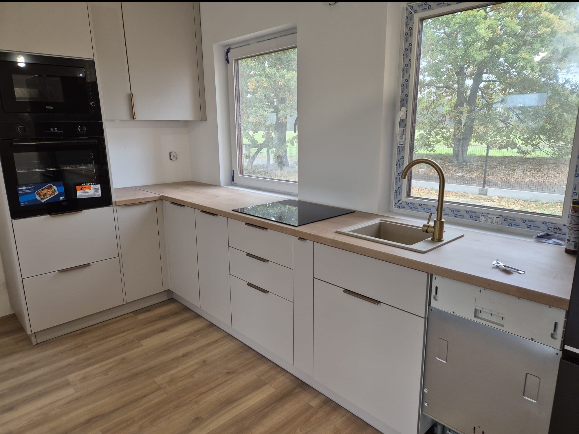 Jasna kuchnia z drewnianym blatem, widok na zewnątrz przez okno. Częściowo zamontowana zmywarka, złota bateria przy zlewie. Nowoczesny design.