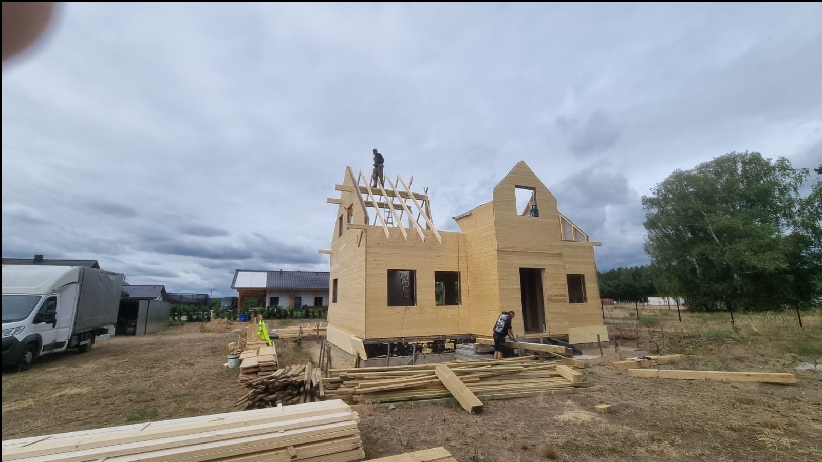 Budowa domu szkieletowego w trakcie realizacji. Widoczna konstrukcja z drewna, pracownicy na dachu i wokół budynku. Na pierwszym planie stos drewna.