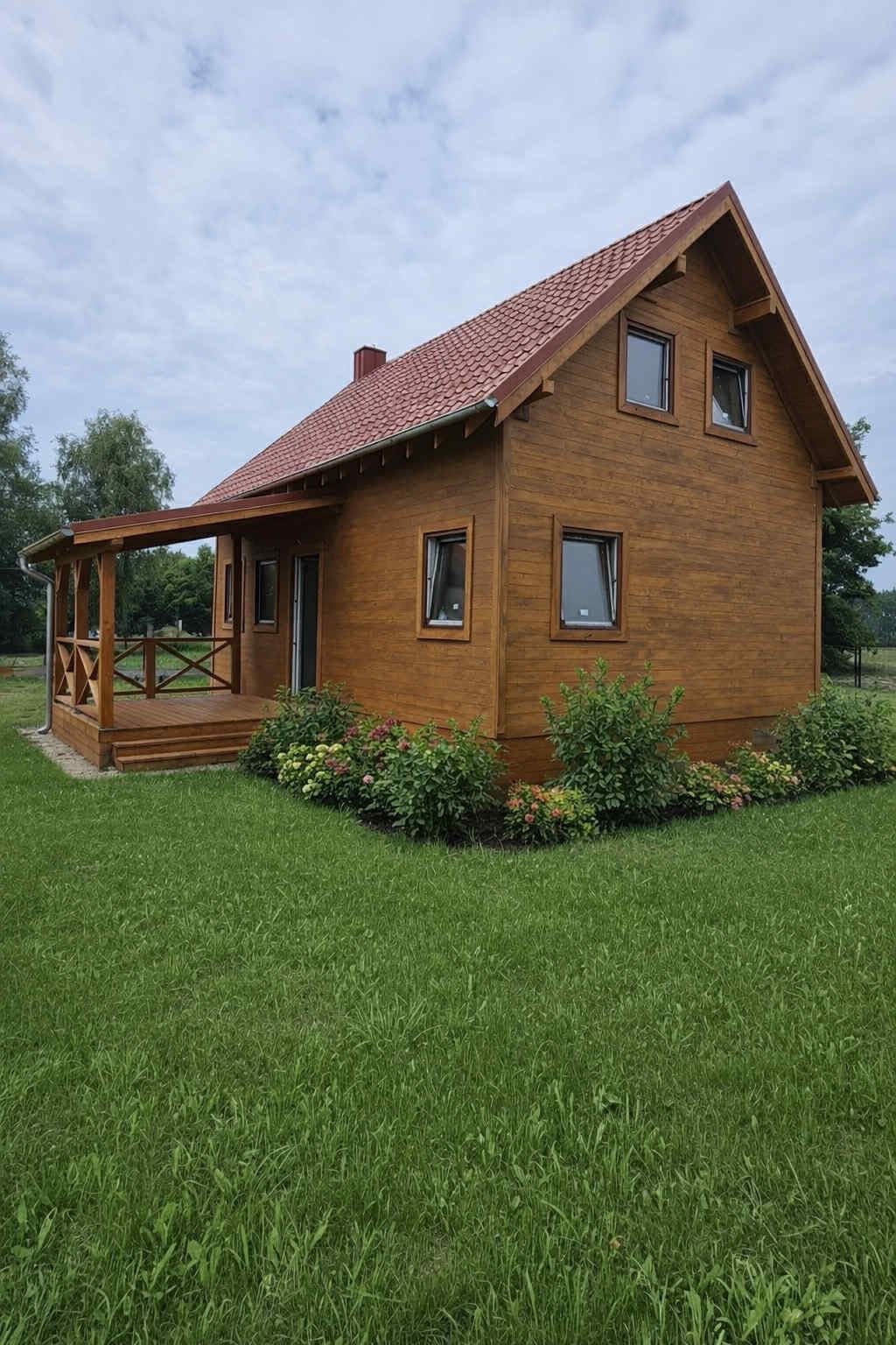 Gotowy, brązowy dom z drewnianą elewacją i czerwonym dachem, otoczony zielonym trawnikiem i krzewami. Widoczny zadaszony taras z drewnianą balustradą.