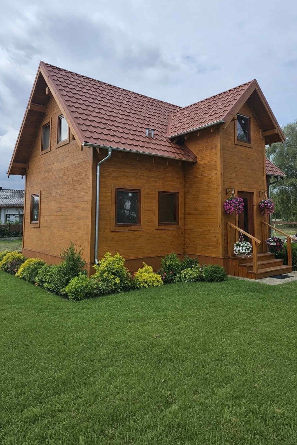 Gotowy, drewniany dom z czerwonym dachem, otoczony zielonym trawnikiem i ozdobnymi krzewami. Widoczne detale wykończenia, takie jak rynny i kwiaty przy wejściu.