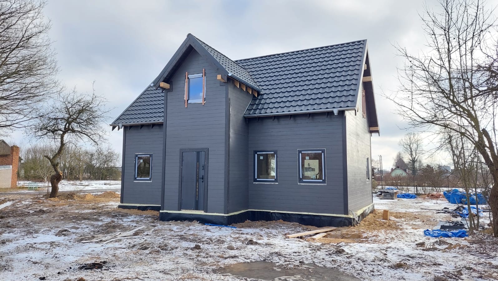 Szary dom z drewnianą elewacją i ciemną dachówką w zimowym krajobrazie. Widoczne drewniane elementy wokół okien i drzwi. Częściowo ośnieżony teren wokół budynku.