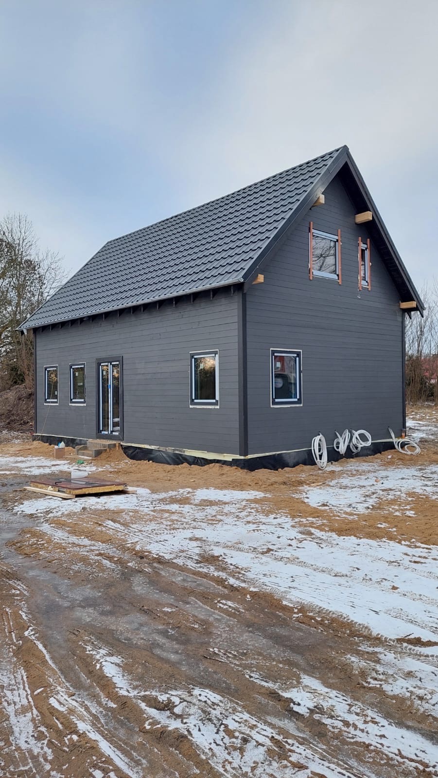 Szary dom z ciemną dachówką w trakcie budowy, widoczne okna i drzwi, teren wokół częściowo pokryty śniegiem, przewody elektryczne na zewnątrz.