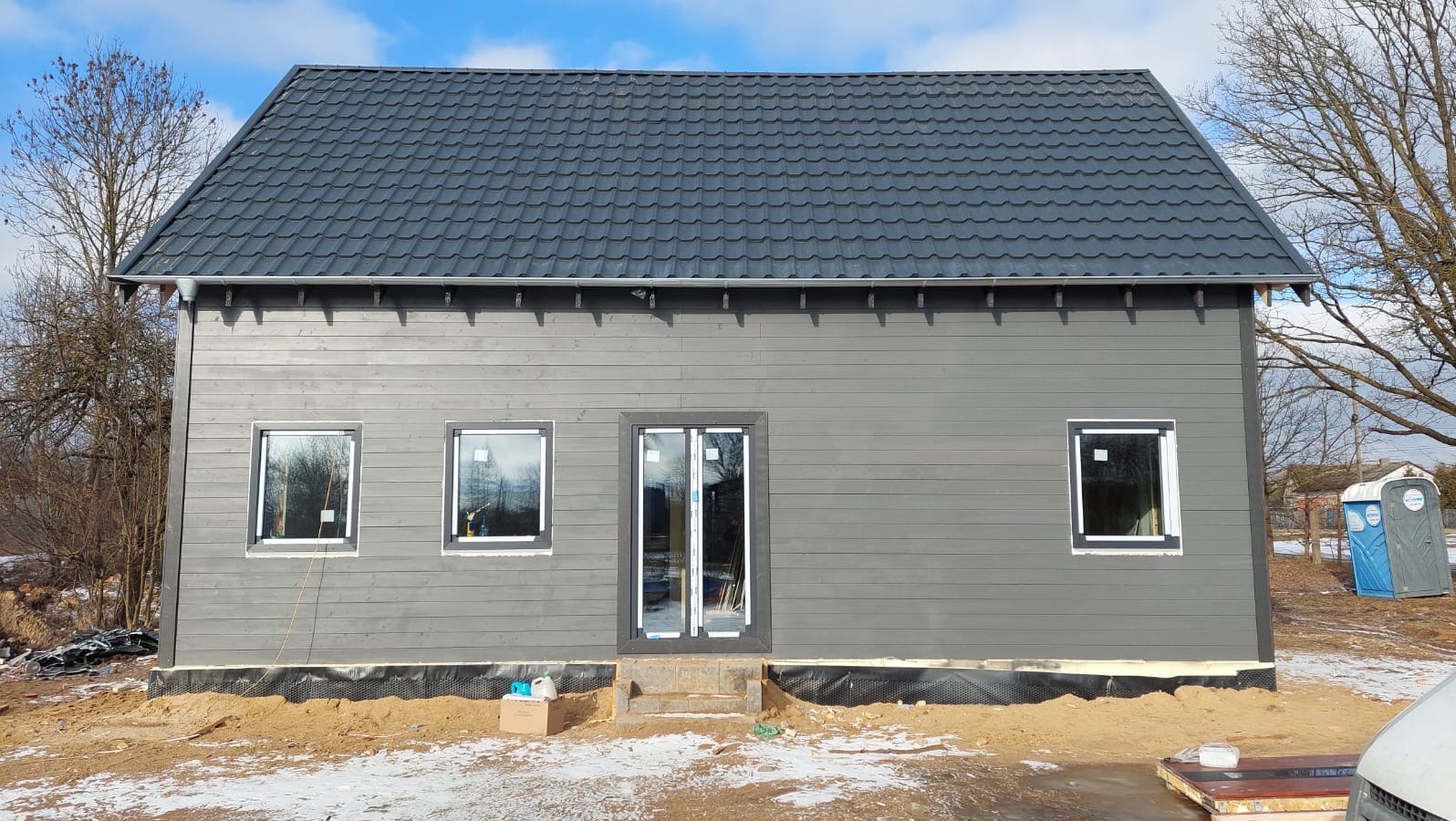 Szary dom szkieletowy z ciemnym dachem z blachodachówki, widoczne okna i drzwi wejściowe. Budynek w trakcie wykończenia, na zewnątrz piasek i przenośna toaleta.