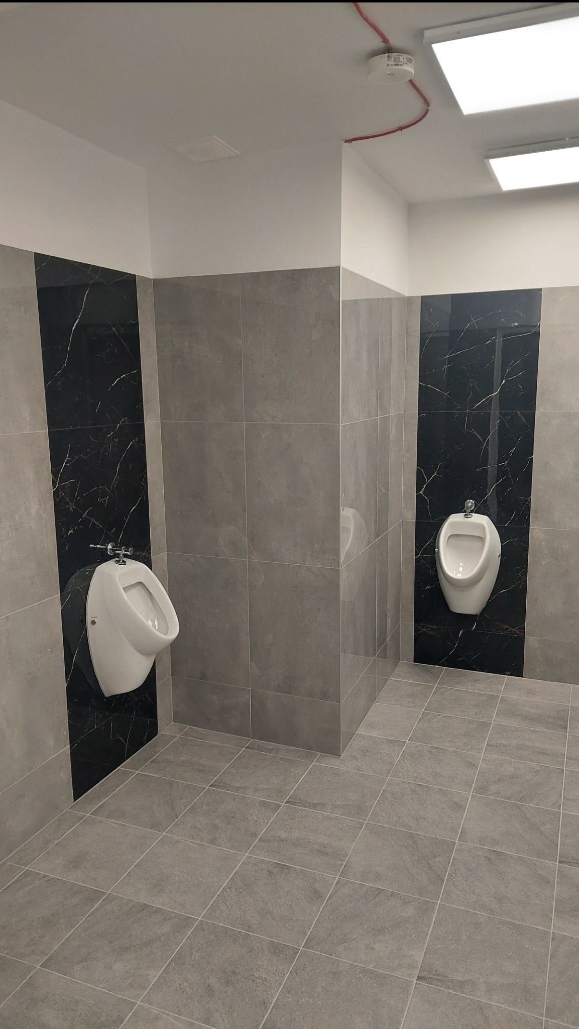 Nowoczesna toaleta publiczna z dwoma białymi pisuarami na ścianach wyłożonych szarymi i czarnymi płytkami imitującymi marmur, szara podłoga z płytek.