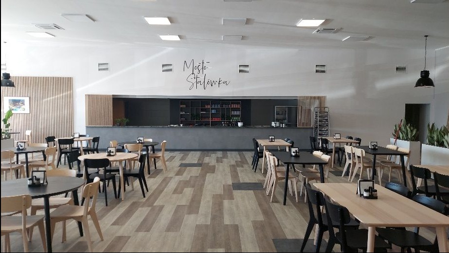 Jasne wnętrze stołówki z drewnianymi panelami, stolikami i krzesłami w odcieniach beżu i czerni. Na ścianie napis 'Mostostalówka'.