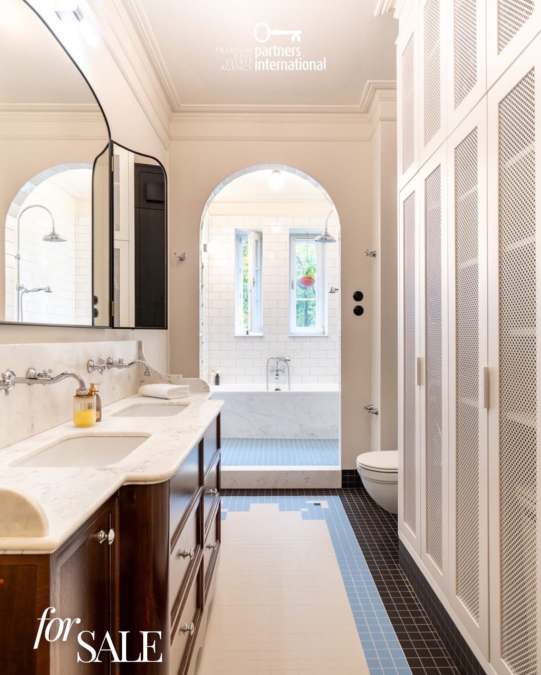 Elegancka łazienka z marmurowym blatem, drewnianymi szafkami i kabiną prysznicową widoczną w lustrze. Na podłodze mozaika w odcieniach bieli, błękitu i czerni. 'For SALE' w rogu.