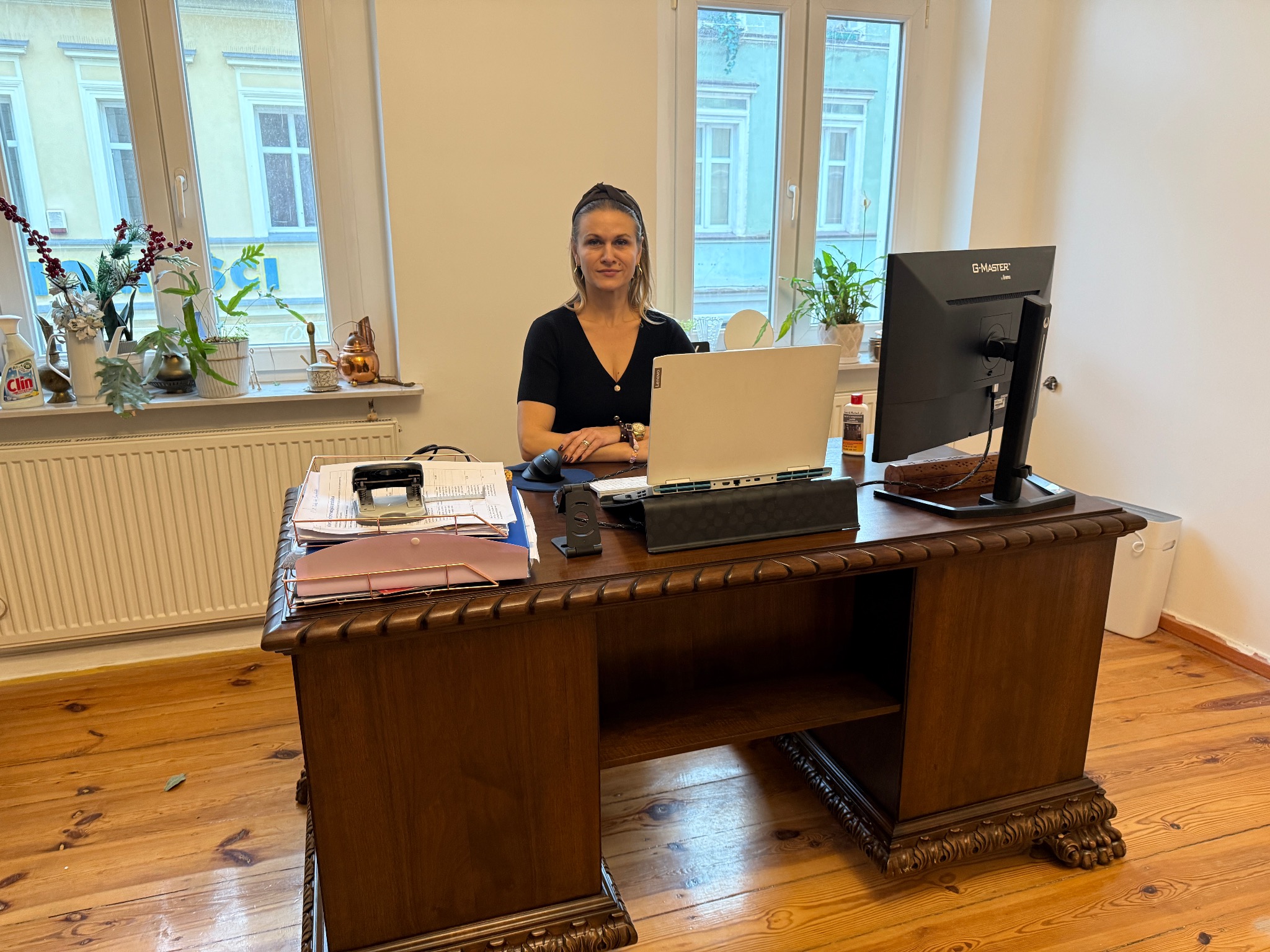 Kobieta siedzi za biurkiem w jasnym biurze. Na biurku laptop, monitor, dokumenty i rośliny. Widoczne okna z roślinami na parapecie. Klasyczny wystrój wnętrza.