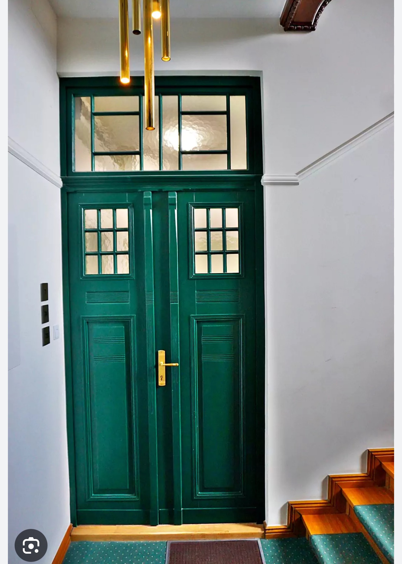 Zielone, dwuskrzydłowe drzwi z szybkami w białej klatce schodowej, oświetlone złotą lampą. Widoczne schody z zieloną wykładziną i brązowy element dekoracyjny pod sufitem.