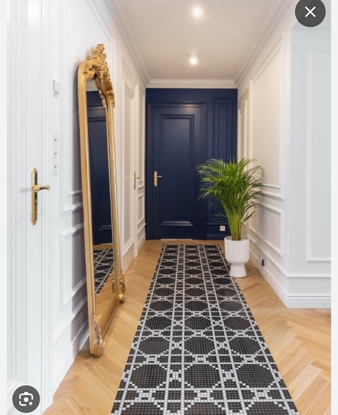 Elegancki korytarz z parkietem w jodełkę, czarno-białym dywanem z mozaiki, wysokim lustrem w złotej ramie i granatowymi drzwiami. W rogu donica z rośliną.