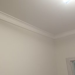 Grzegbud - Wykończony narożnik pokoju z listwą dekoracyjną pod sufitem i widocznym fragmentem ościeżnicy drzwiowej. Ściany pomalowane na jasny, neutralny kolor.