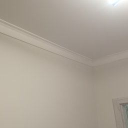 Grzegbud - Wykończony narożnik pokoju z listwą przysufitową w kolorze białym. Widoczny fragment oświetlenia sufitowego i górna część ościeżnicy drzwiowej.