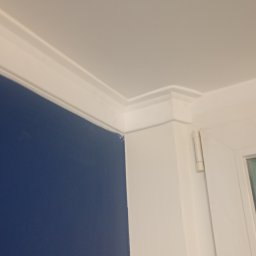 Grzegbud - Detal wykończenia wnętrza: białe listwy przysufitowe zdobiące połączenie niebieskiej ściany i białego sufitu, widoczny fragment białej ramy okiennej.