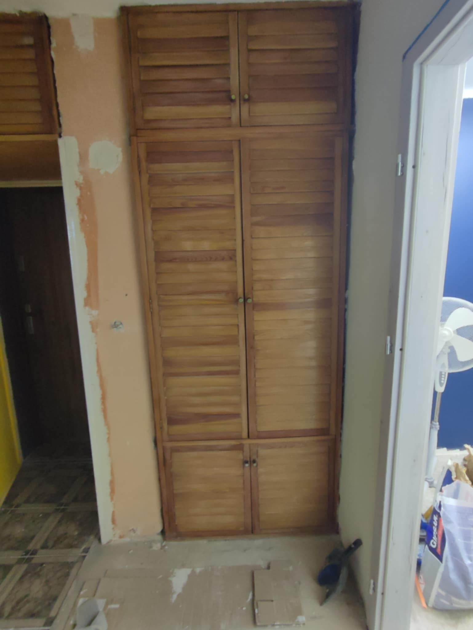 Drewniana szafa z lamelowymi drzwiami w trakcie montażu, widoczne niedokończone ściany i podłoga. Fragment wentylatora w tle. Ujęcie z bliska, naturalne światło.