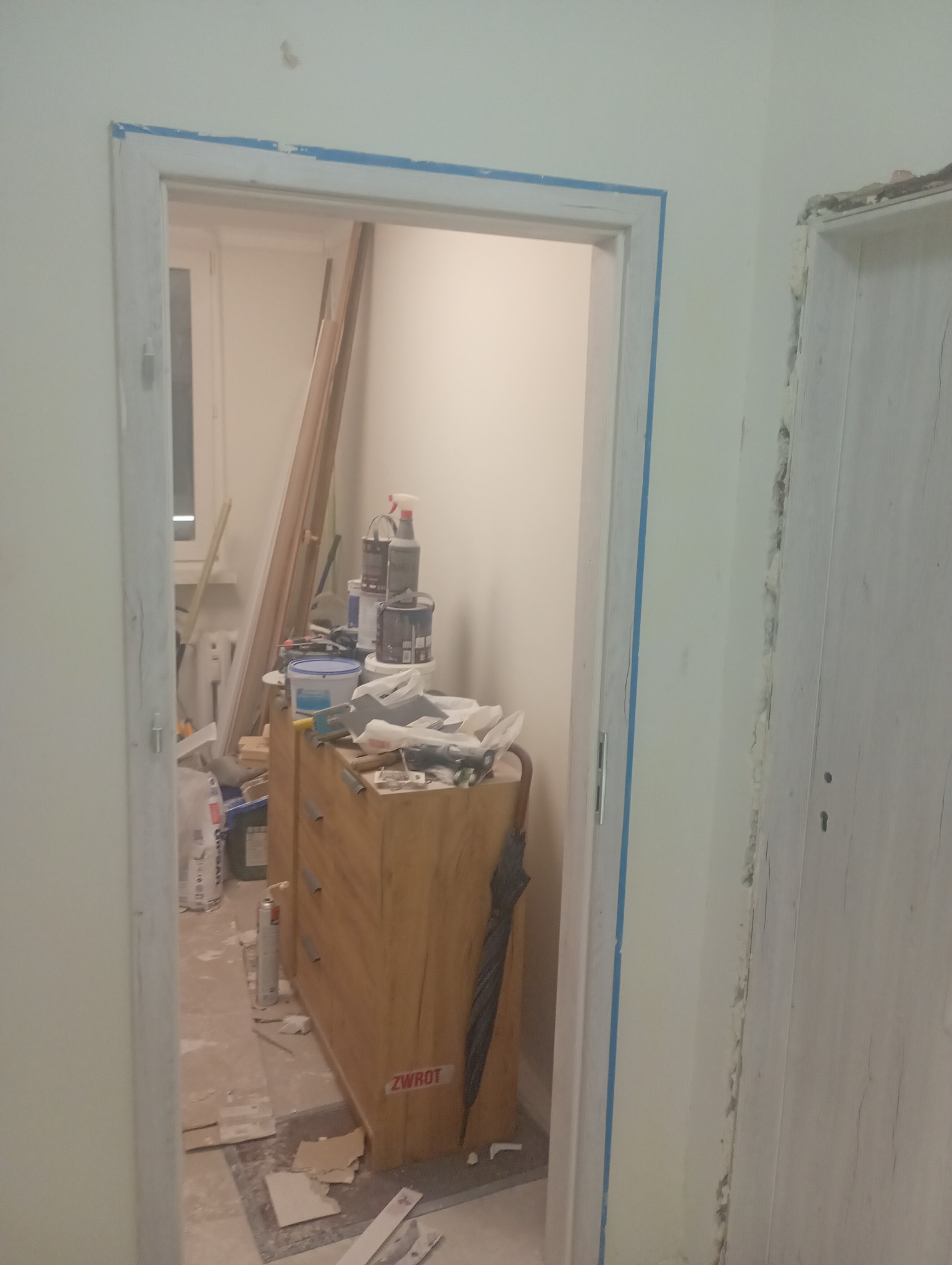 Remont mieszkania: widok na pokój przez ościeżnicę z niebieską taśmą malarską. W tle farby, narzędzia i komoda. Podłoga w trakcie prac wykończeniowych.