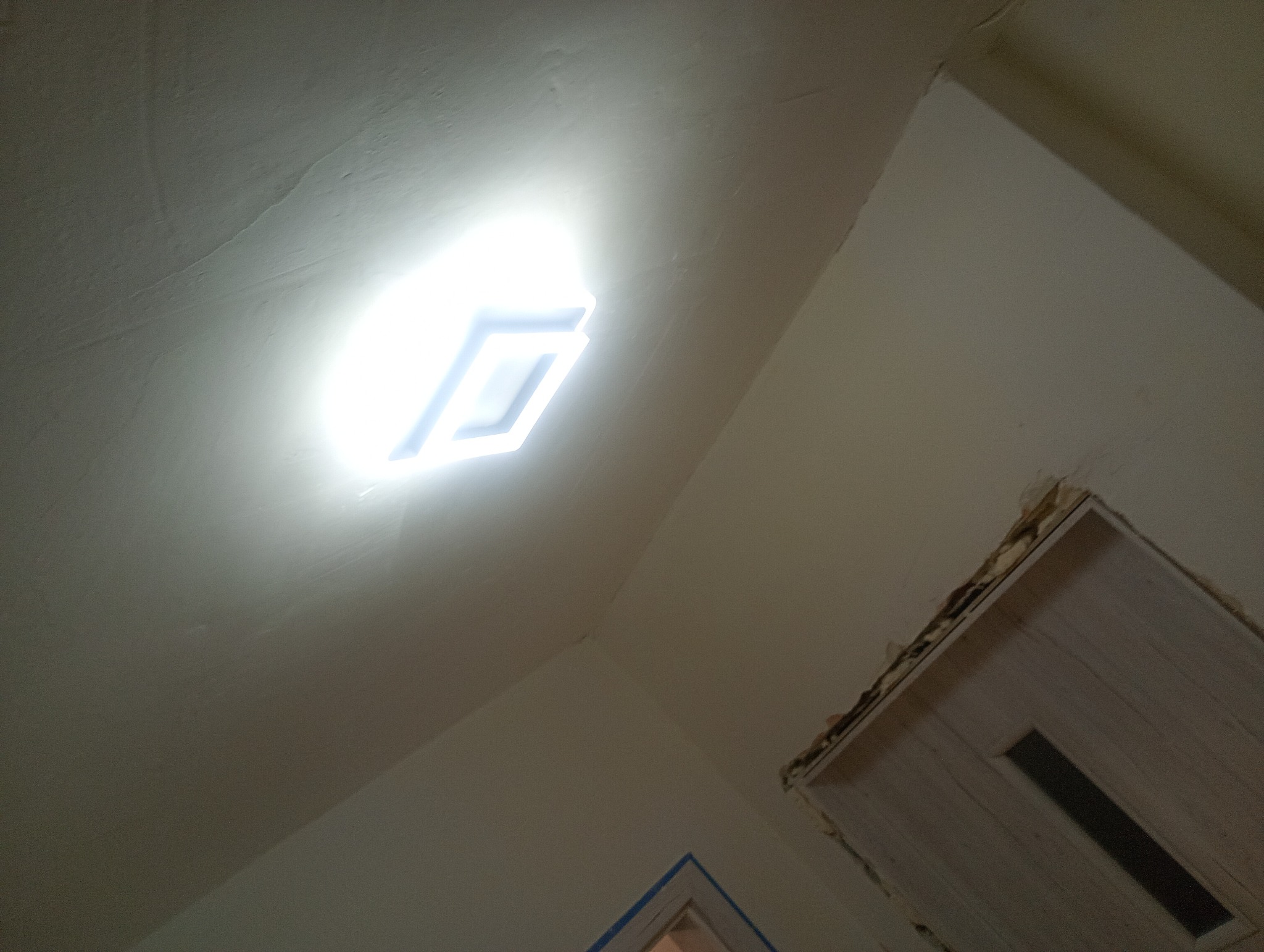 Nisko kątowe ujęcie sufitu z zamontowanym, świecącym panelem LED i fragmentem ościeżnicy drzwiowej w trakcie prac wykończeniowych.