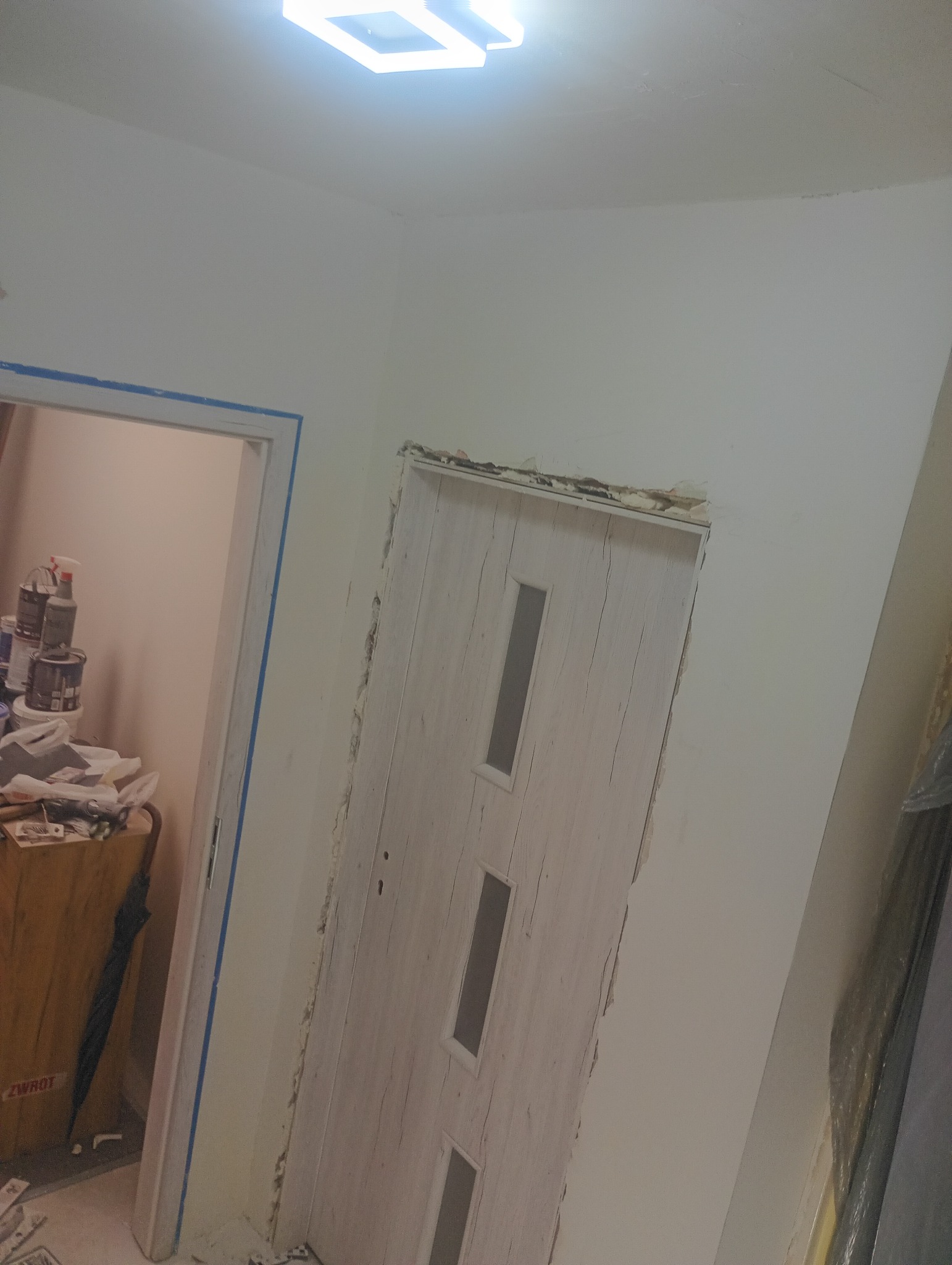 Montaż ościeżnicy drzwiowej z widocznymi szczelinami i taśmą malarską, w tle fragment pomieszczenia z narzędziami i farbami. Prace wykończeniowe w trakcie.