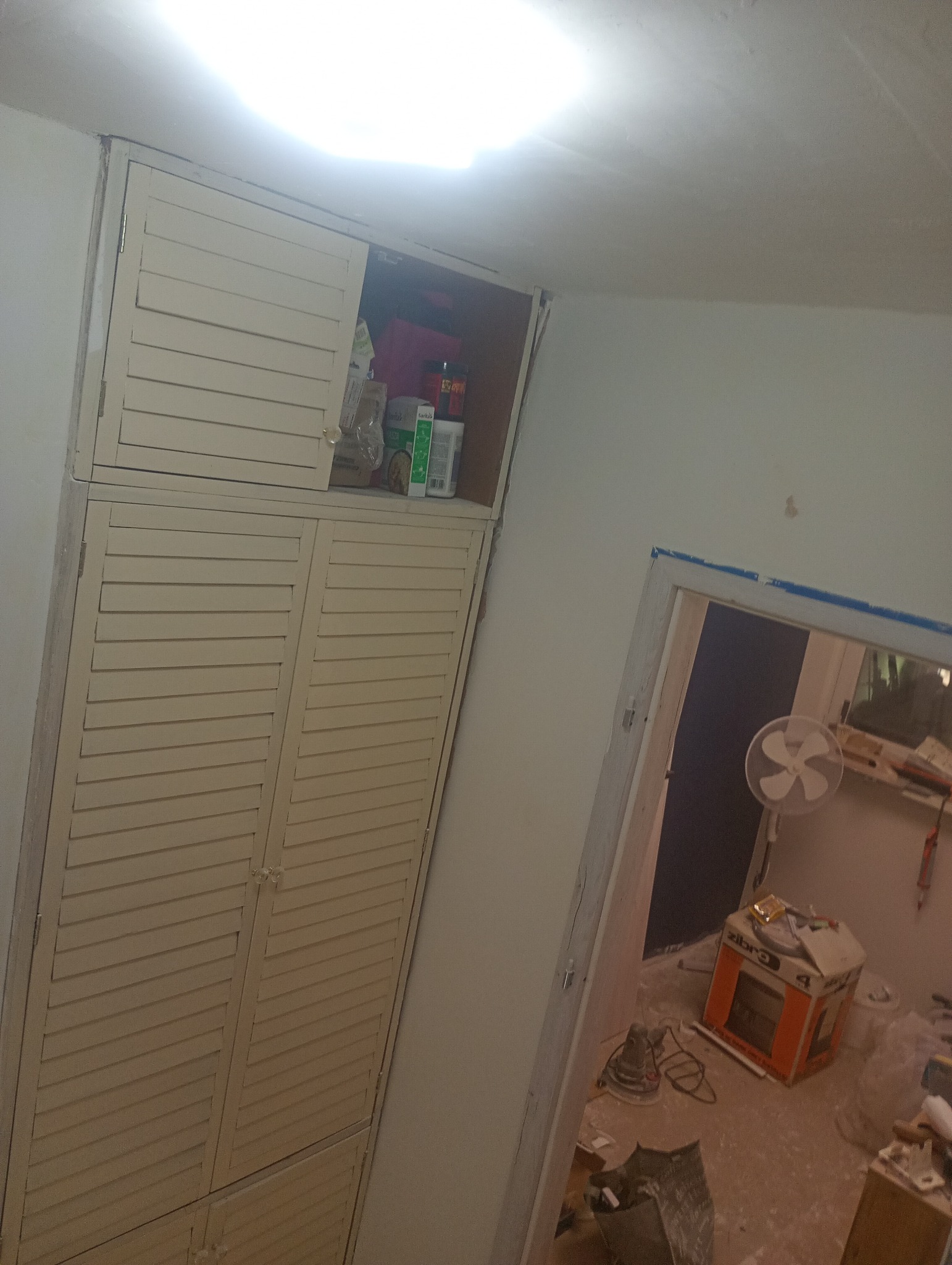 Biała szafa z lamelowymi drzwiami w trakcie malowania, widoczne narzędzia i materiały malarskie, wnętrze w trakcie remontu.