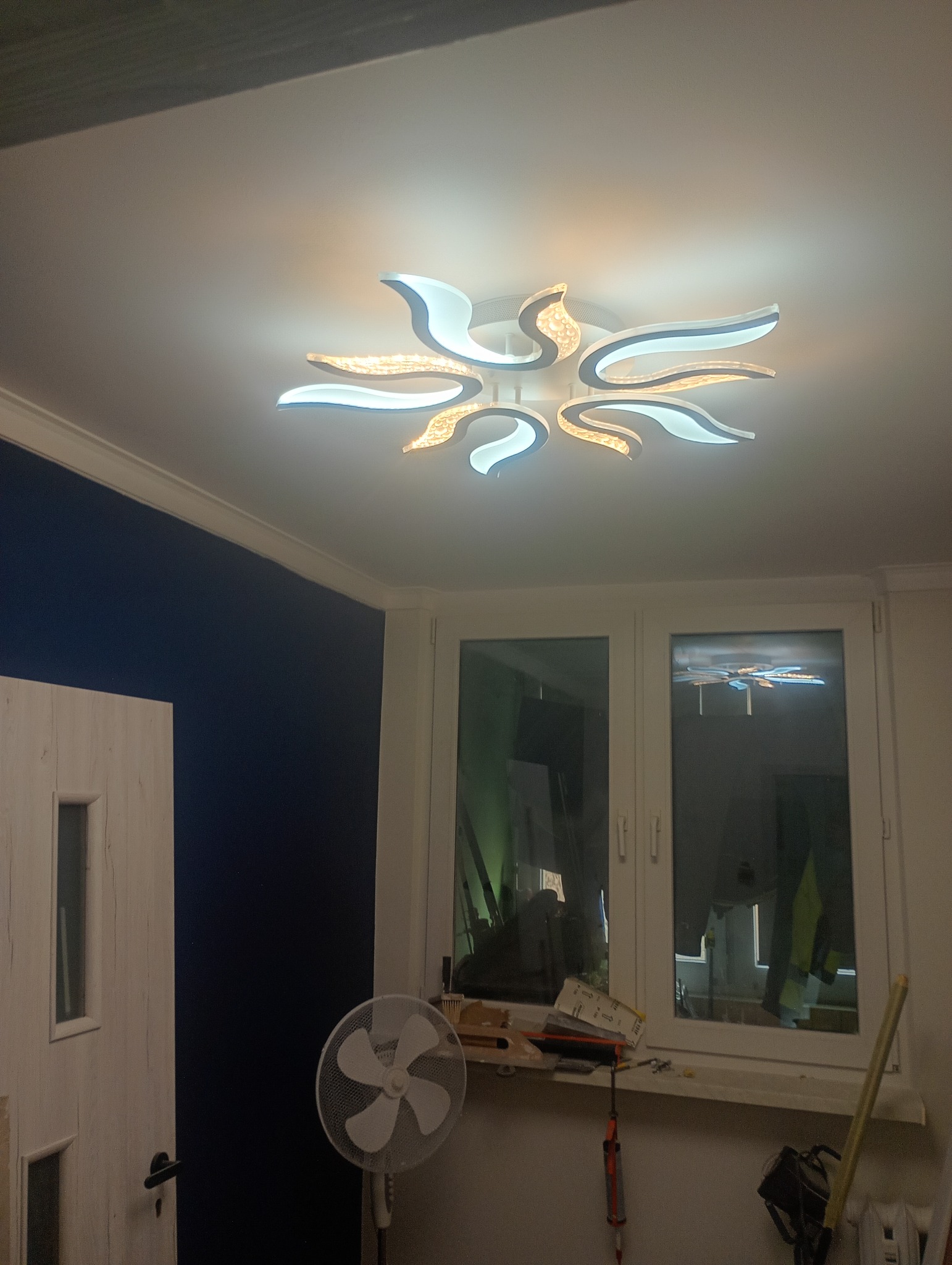 Nowoczesna lampa sufitowa w kształcie kwiatu, oświetlająca pokój z niebieską ścianą i białymi oknami. Widoczny wentylator i narzędzia na parapecie.