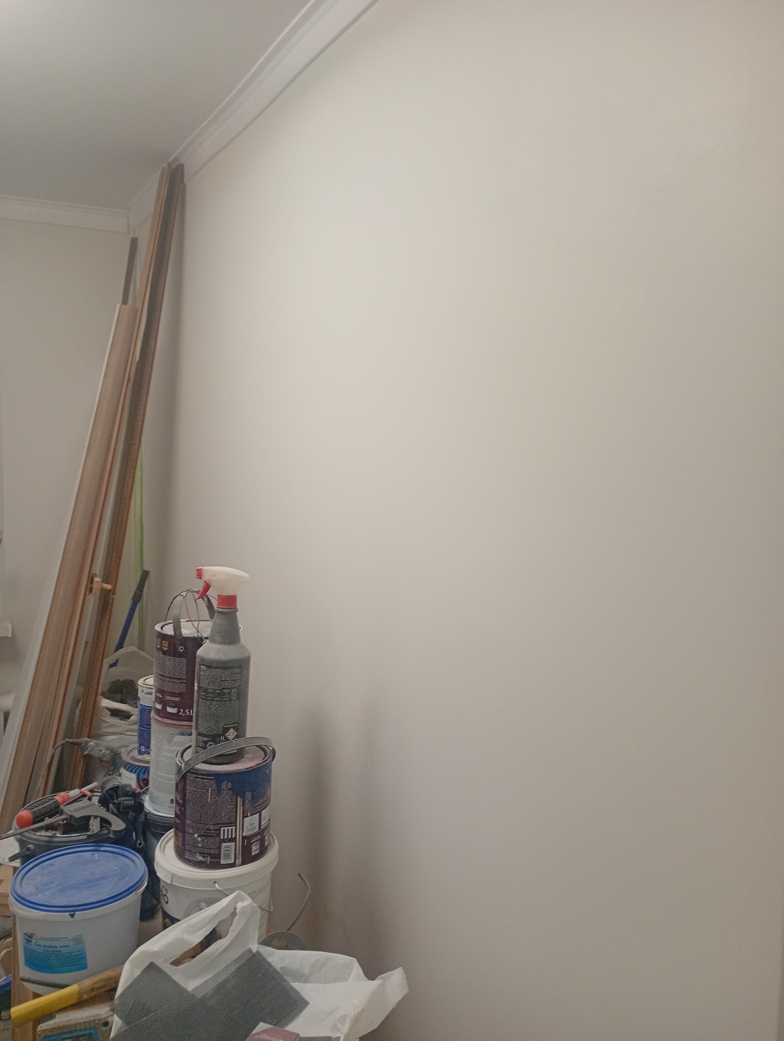 Ściana po malowaniu z widocznymi narzędziami malarskimi i puszkami farb, listwy przypodłogowe oparte o ścianę. Przygotowania do dalszych prac wykończeniowych.
