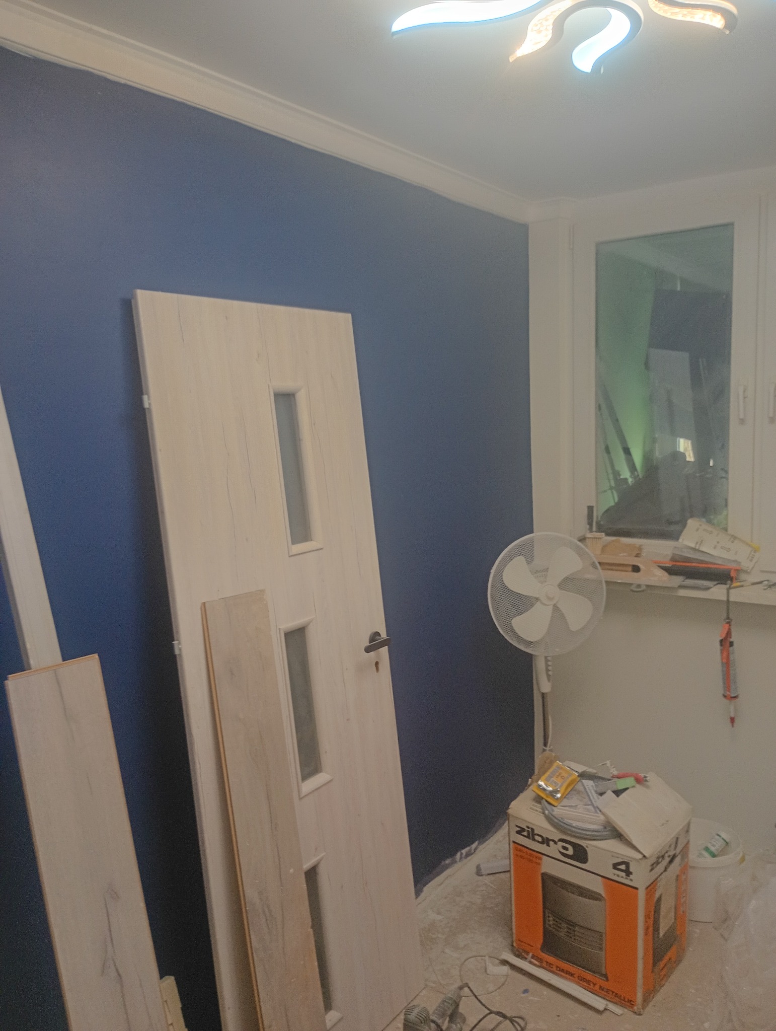 Wnętrze w trakcie remontu: niebieska ściana, białe drzwi z szybkami, panele podłogowe, wentylator i karton. Widoczne narzędzia i akcesoria remontowe.