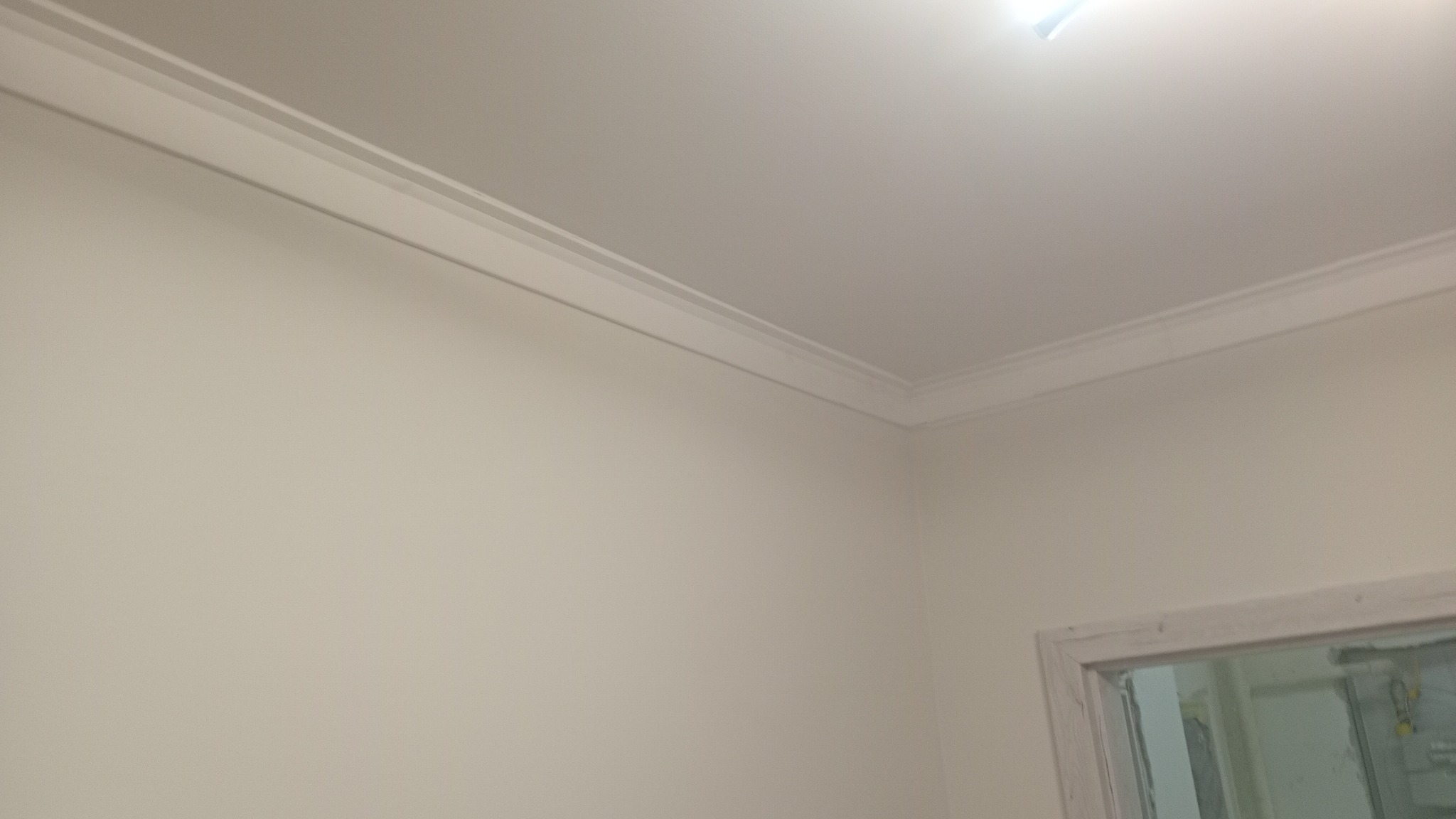 Wykończony narożnik pokoju z listwą dekoracyjną pod sufitem i widocznym fragmentem ościeżnicy drzwiowej. Ściany pomalowane na jasny, neutralny kolor.