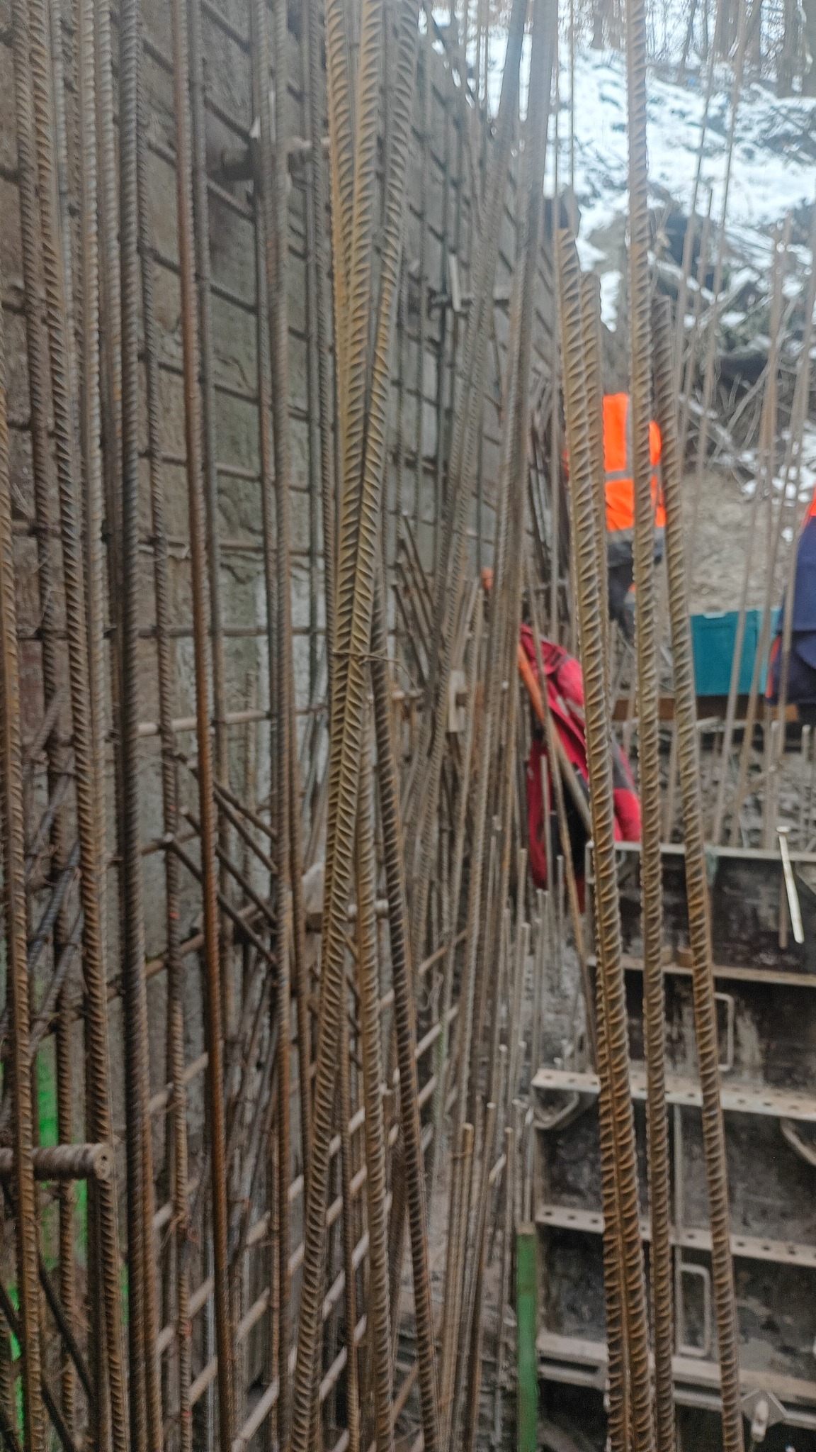 Zbrojenie pionowe i poziome w szalunku, widoczne pręty żebrowane, fragmenty form i sylwetki pracowników w tle. Surowy etap budowy, detale konstrukcyjne.