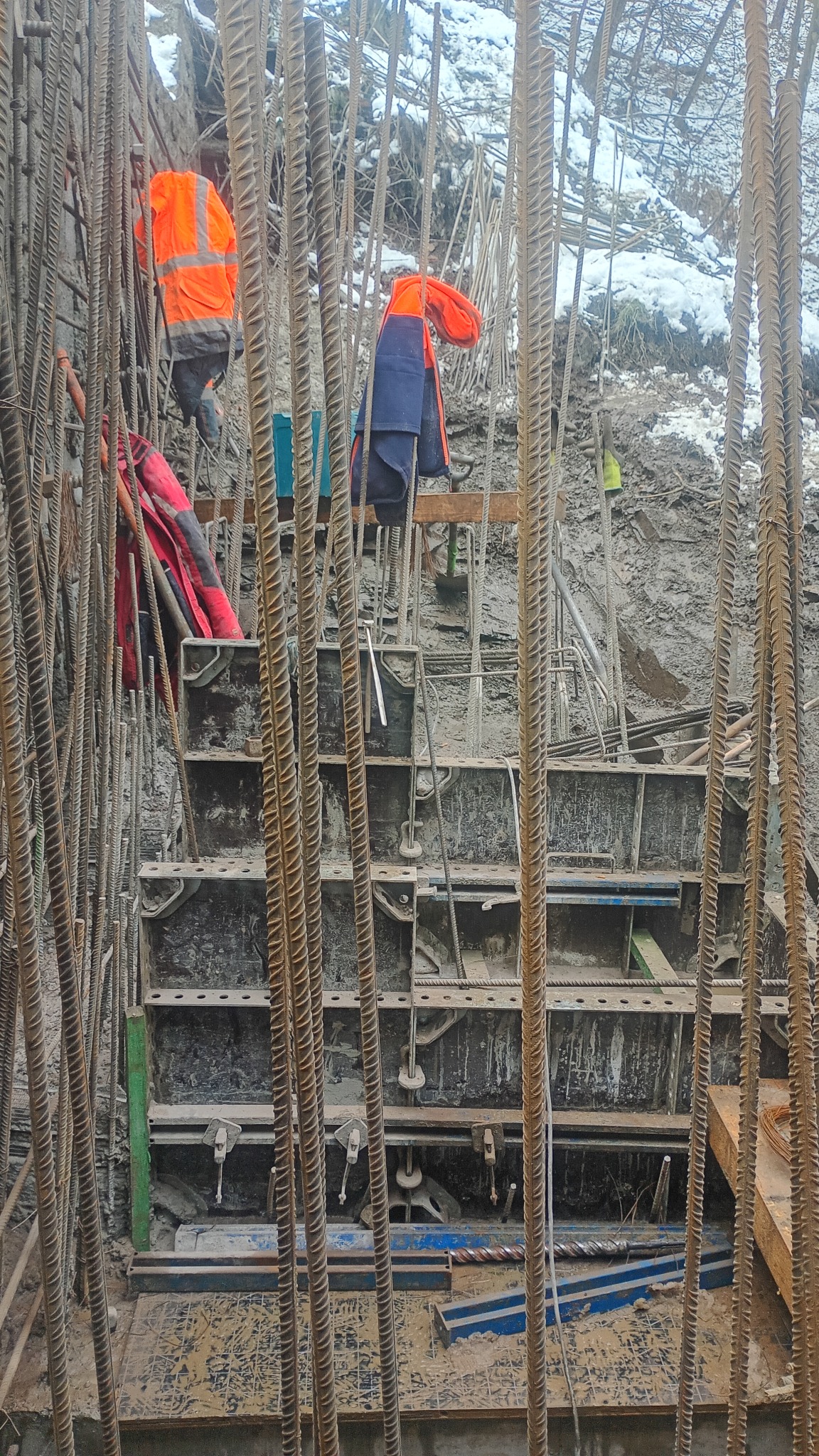 Zbrojenie fundamentów pod budynek w trakcie prac. Widoczne stalowe pręty, szalunki i pracownicy w odblaskowych ubraniach. Prace ziemne w zimowych warunkach.