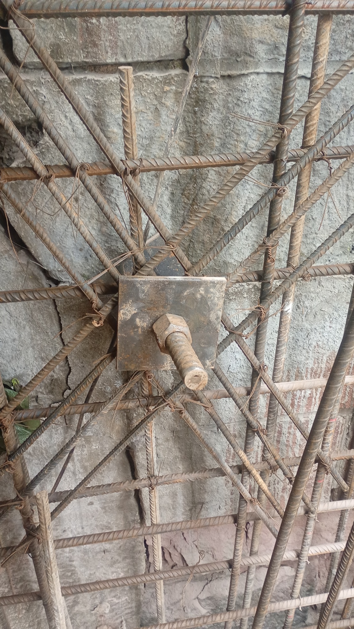 Zbrojenie ściany z prętów stalowych, połączonych drutem, z kwadratową płytką i śrubą na środku, na tle szarej, surowej ściany z betonu lub kamienia.