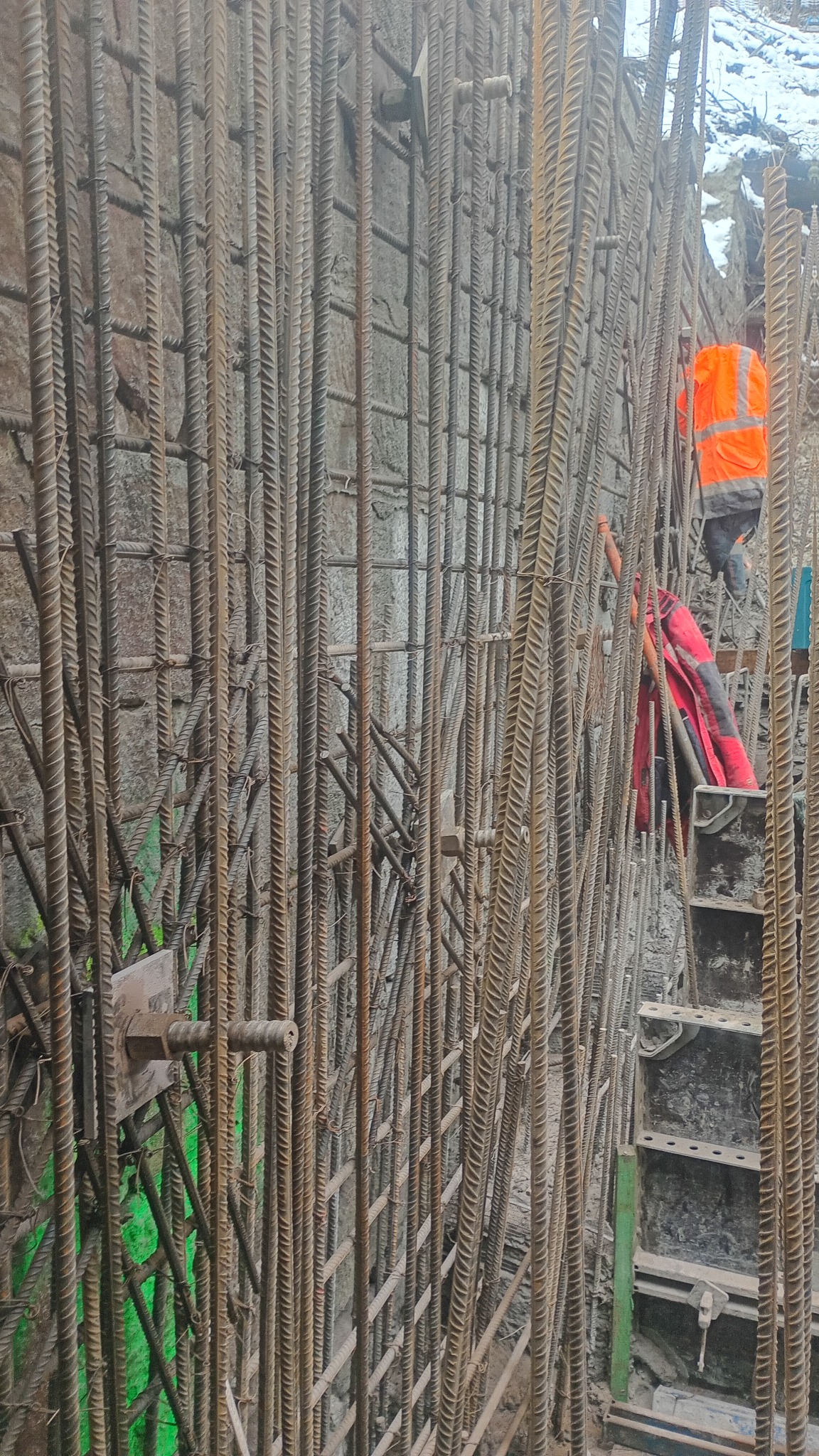 Zbrojenie ściany z betonu z widocznymi prętami i elementami szalunku. W tle pracownik w odblaskowej kamizelce. Etap budowy z bliska.