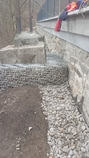 Gabiony z kamienia na zboczu, zabezpieczenie skarpy przed osuwaniem. Metalowa siatka wypełniona kamieniami, surowy beton i ziemia. Prace ziemne i umocnienia.